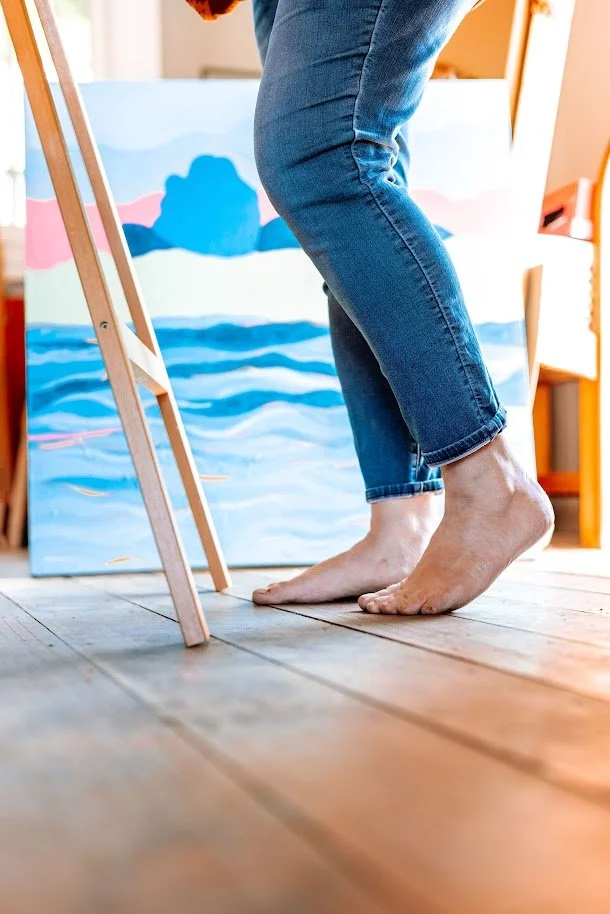 A person with rolled-up jeans is barefoot inside an art studio, standing on wooden floors near a partially visible easel and a painting of a landscape in the background.