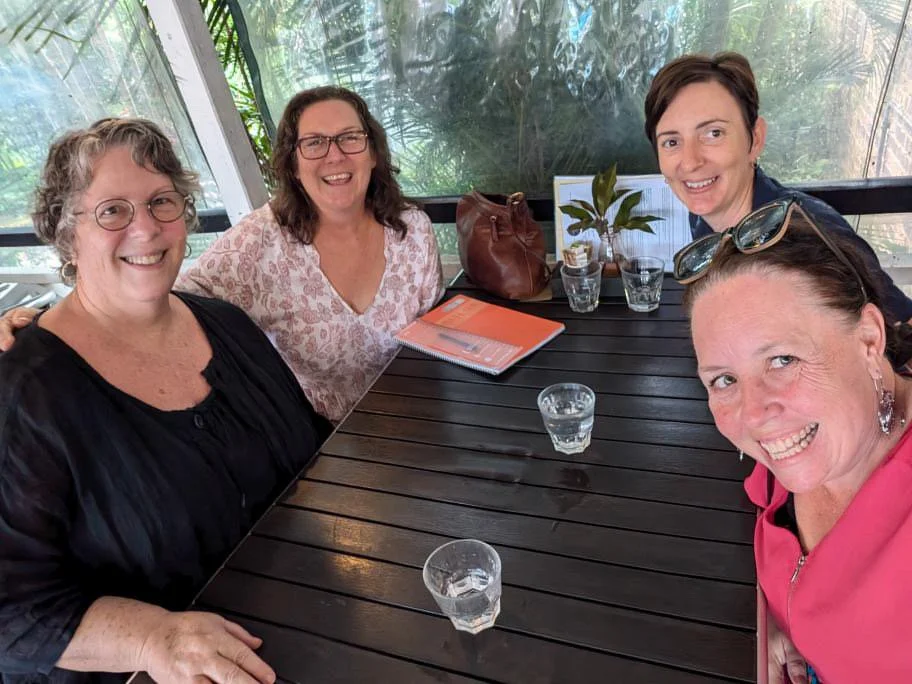 Four women sitting at a black outdoor table with glasses of water, smiling, with a plant, notebooks, and handbags on the table, in a lush, green patio setting.