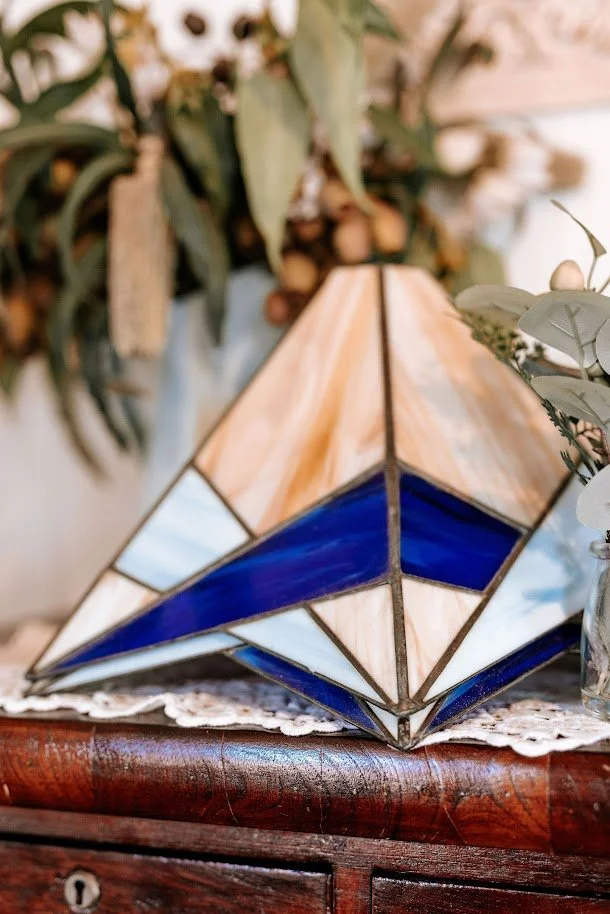 A stained glass geometric pyramid-shaped decorative object with shades of blue, beige, and clear glass, placed on a wooden surface with greenery in the background.