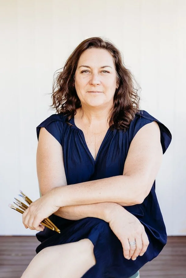 A woman with shoulder-length brown hair sitting on a stool against a plain white background, wearing a navy blue dress, holding paintbrushes in her left hand, with her arms crossed and a slight smile.