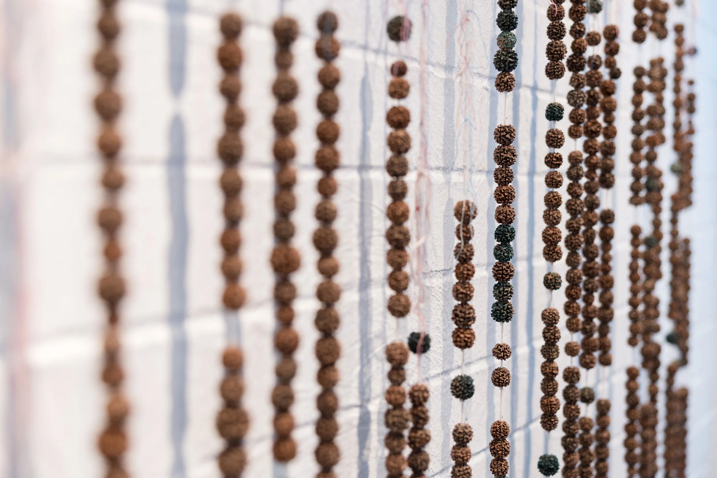Close-up of strings of small, round, brown, green, and black beads hanging vertically against a white brick wall.