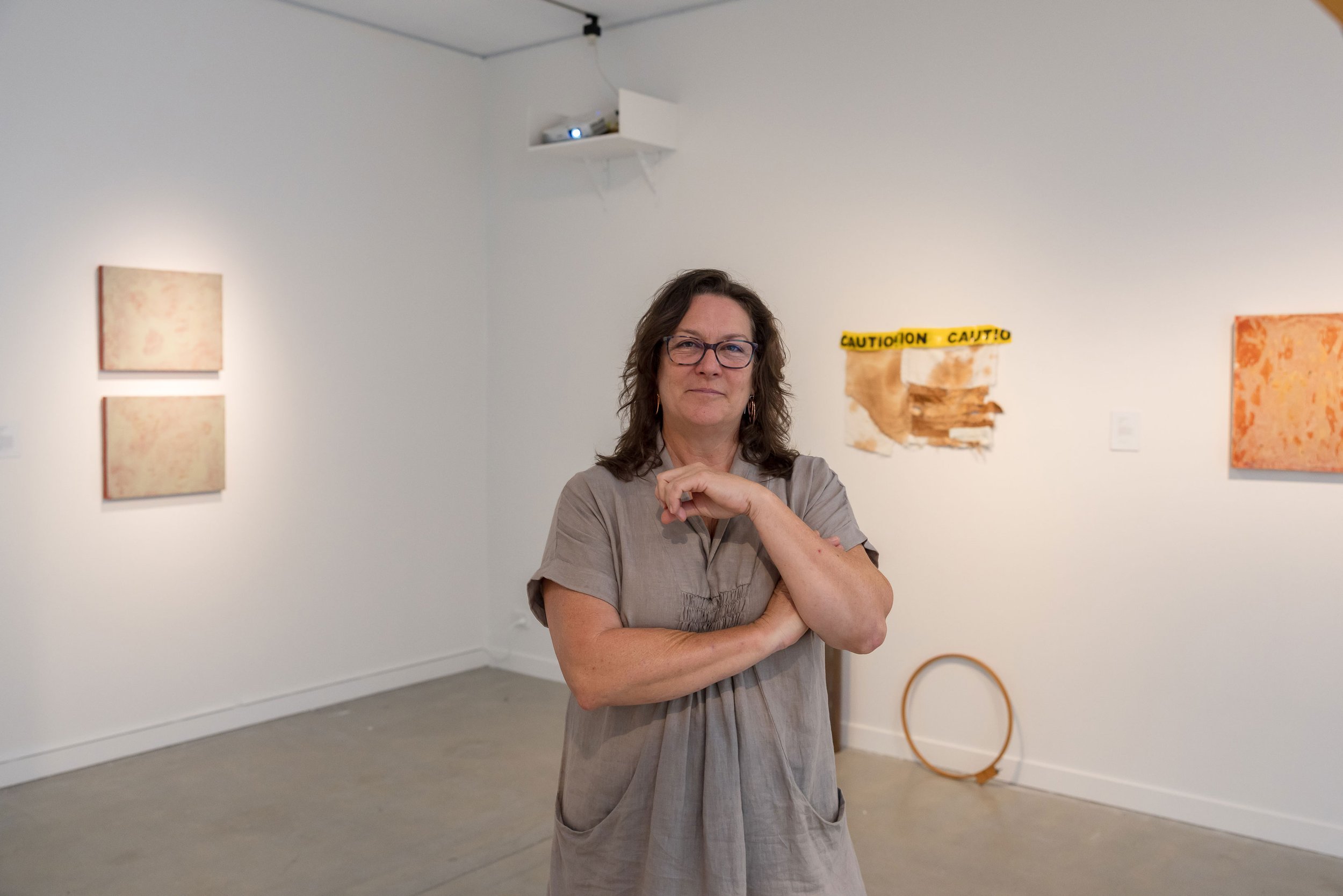 A woman with glasses and curly brown hair stands with arms crossed in an art gallery, surrounded by abstract paintings on white walls.