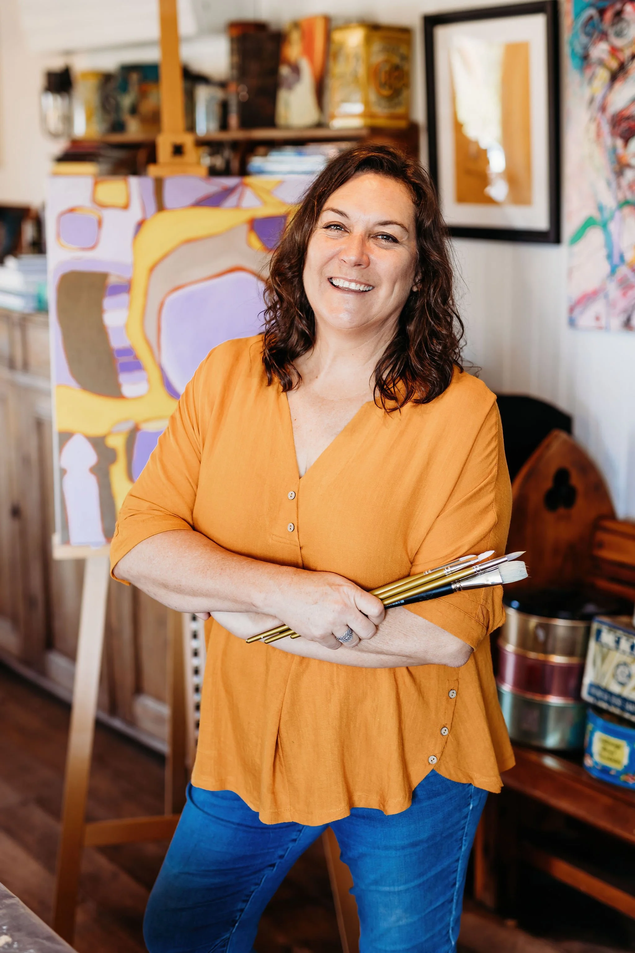 A woman with curly brown hair wearing an orange blouse and blue jeans, holding paintbrushes in an art studio, smiling at the camera.
