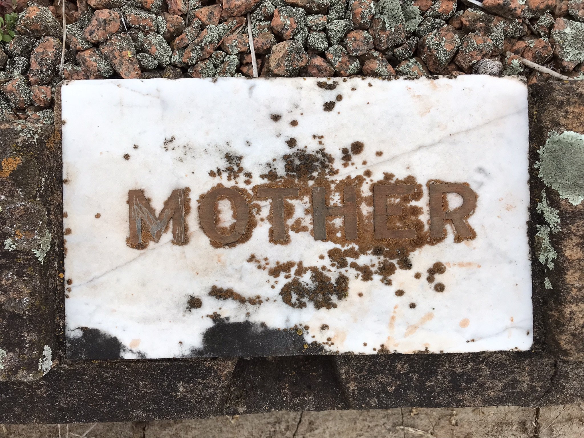A white concrete tablet with the word "MOTHER" etchedinto it, placed on a border of stones and dirt, with some dirt and lichen on the marble surface.