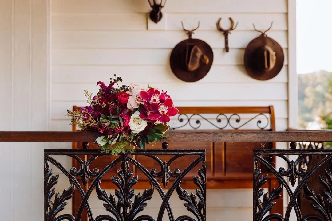 A bouquet of pink, white, and purple flowers resting on a black wrought iron railing of a porch, with a wooden bench and mounted antlers on a white wall in the background.