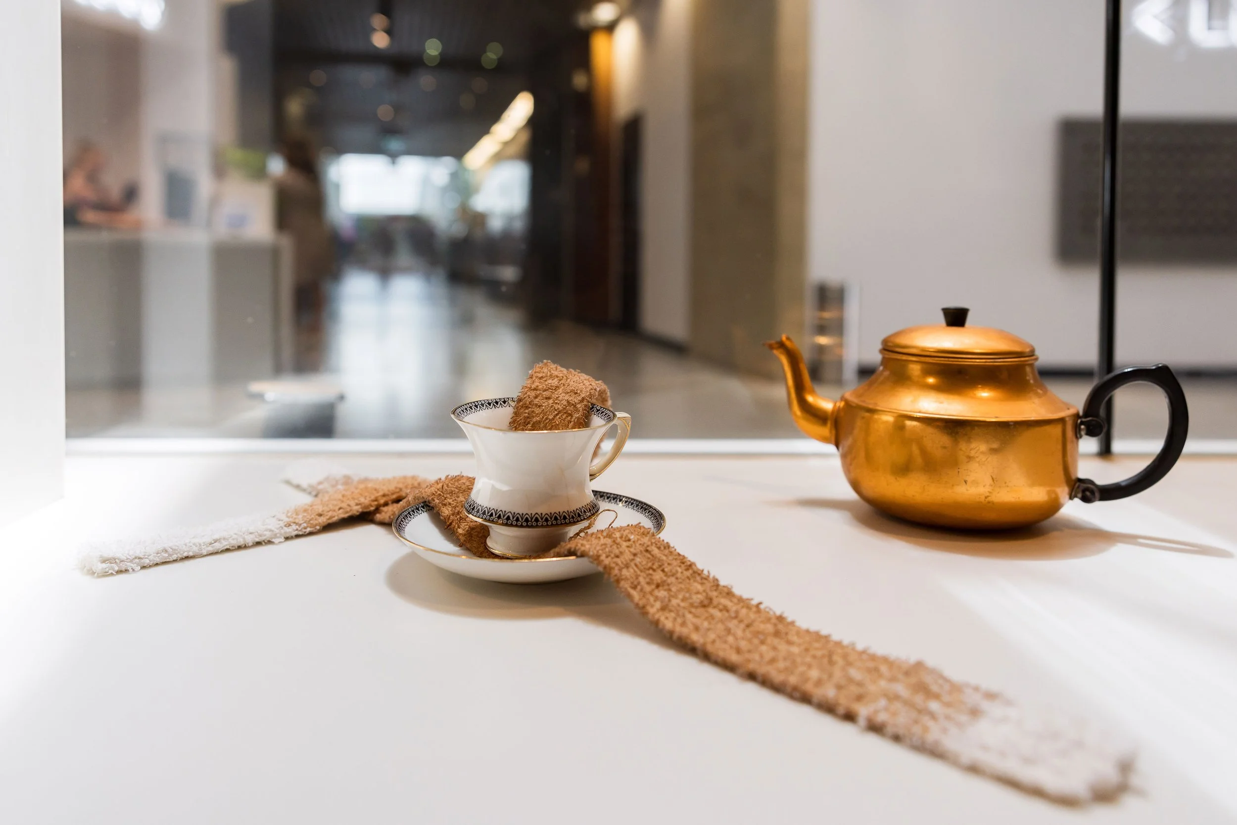 A gold teapot and a white teacup with a saucer, both placed on a white surface in front of a glass window. The teacup contains a brown, textured sponge. There are two textured strips, one white and one brown, laid out on the surface. The background s