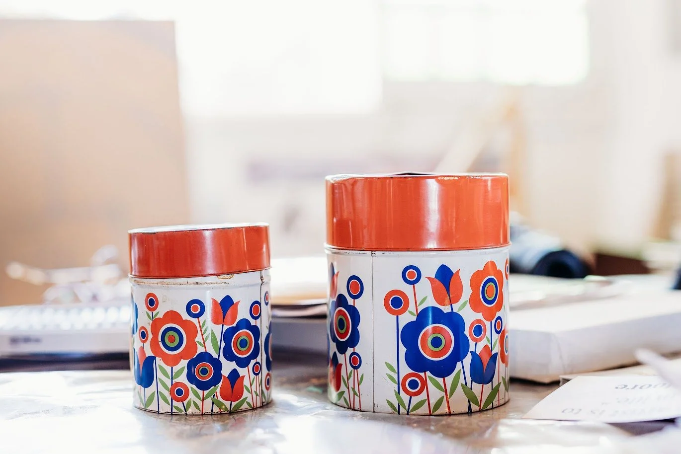 Two cylindrical tins with orange lids and a floral design in red, blue, and green situated on a table with papers and other objects in the background.