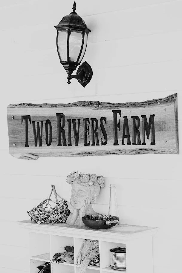 A black and white photo of a wall-mounted lantern, a wooden sign reading 'Two Rivers Farm', and a shelf with decorative items including a bust with a flower crown, a glass bottle, a bowl, and miscellaneous objects.