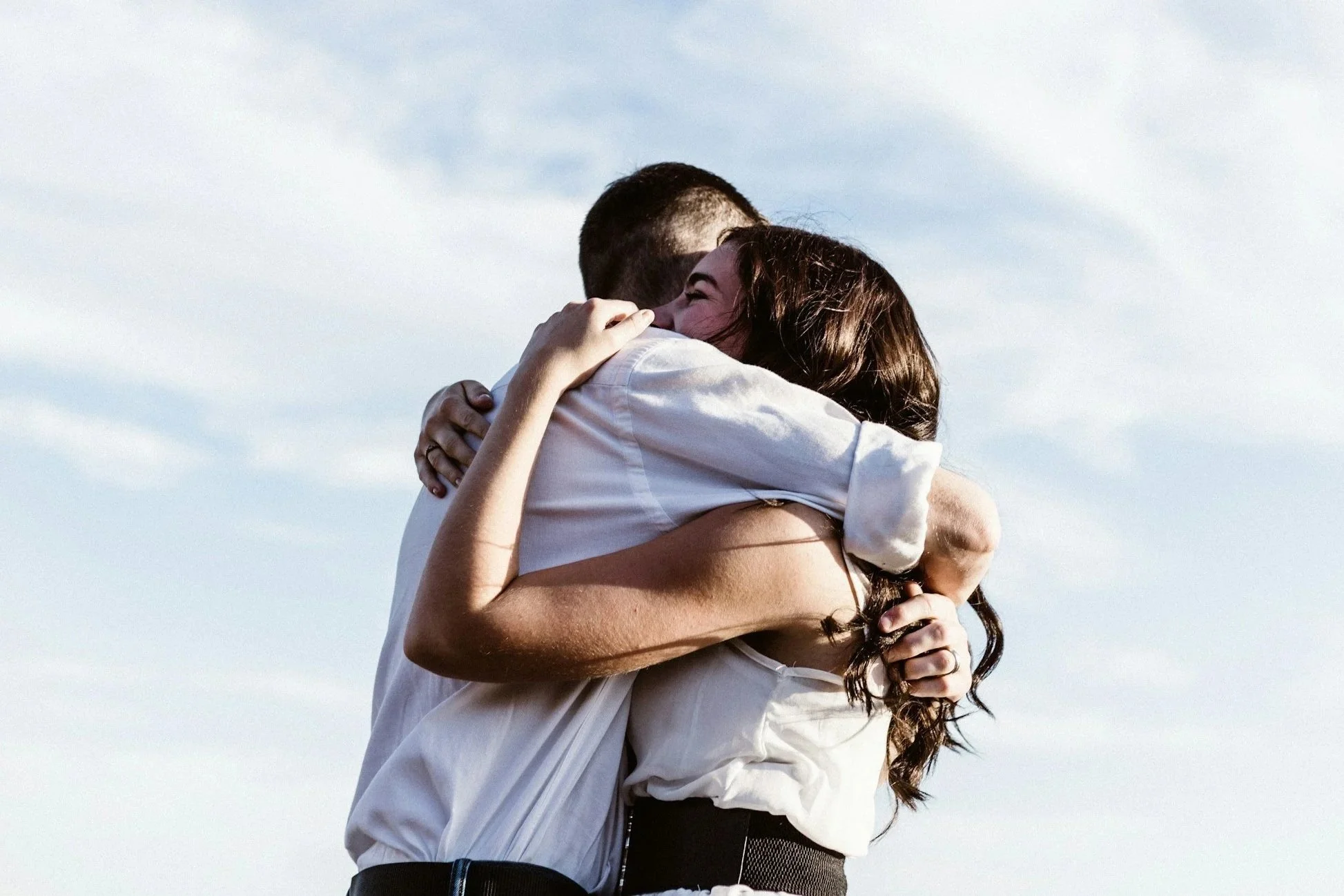 A couple embracing outdoors, sky in the background, the woman smiling, both hugging tightly.