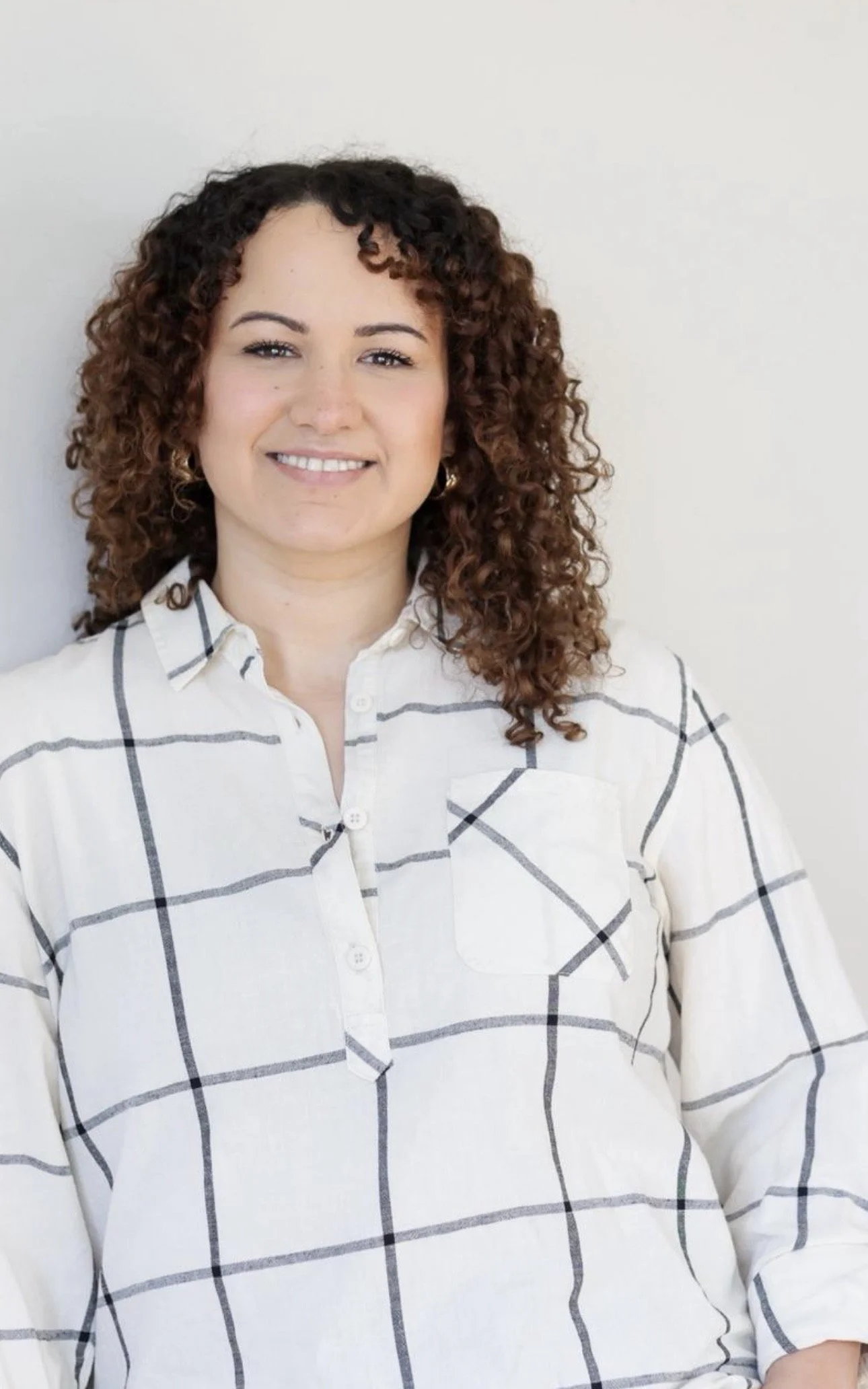 A woman with curly hair wearing a white plaid shirt, smiling, standing against a plain white background.