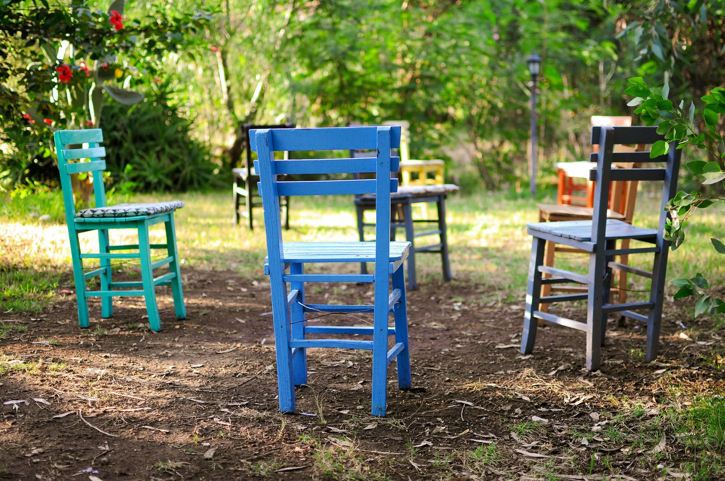 Colorful wooden chairs in blue, turquoise, black, orange, and white are arranged outdoors on a dirt patch amidst green foliage and trees, with some flowers visible.