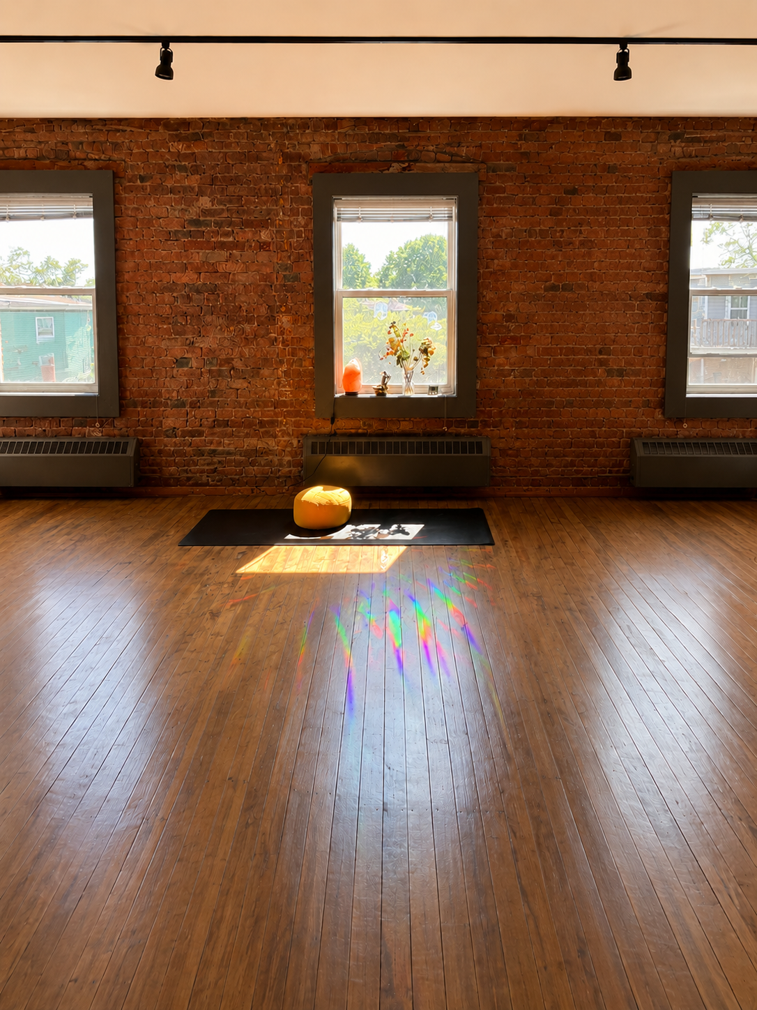Empty room with wooden floor, brick wall, three windows with blinds, a black yoga mat, a yellow meditation cushion, and sunlight creating rainbow reflections on the floor.