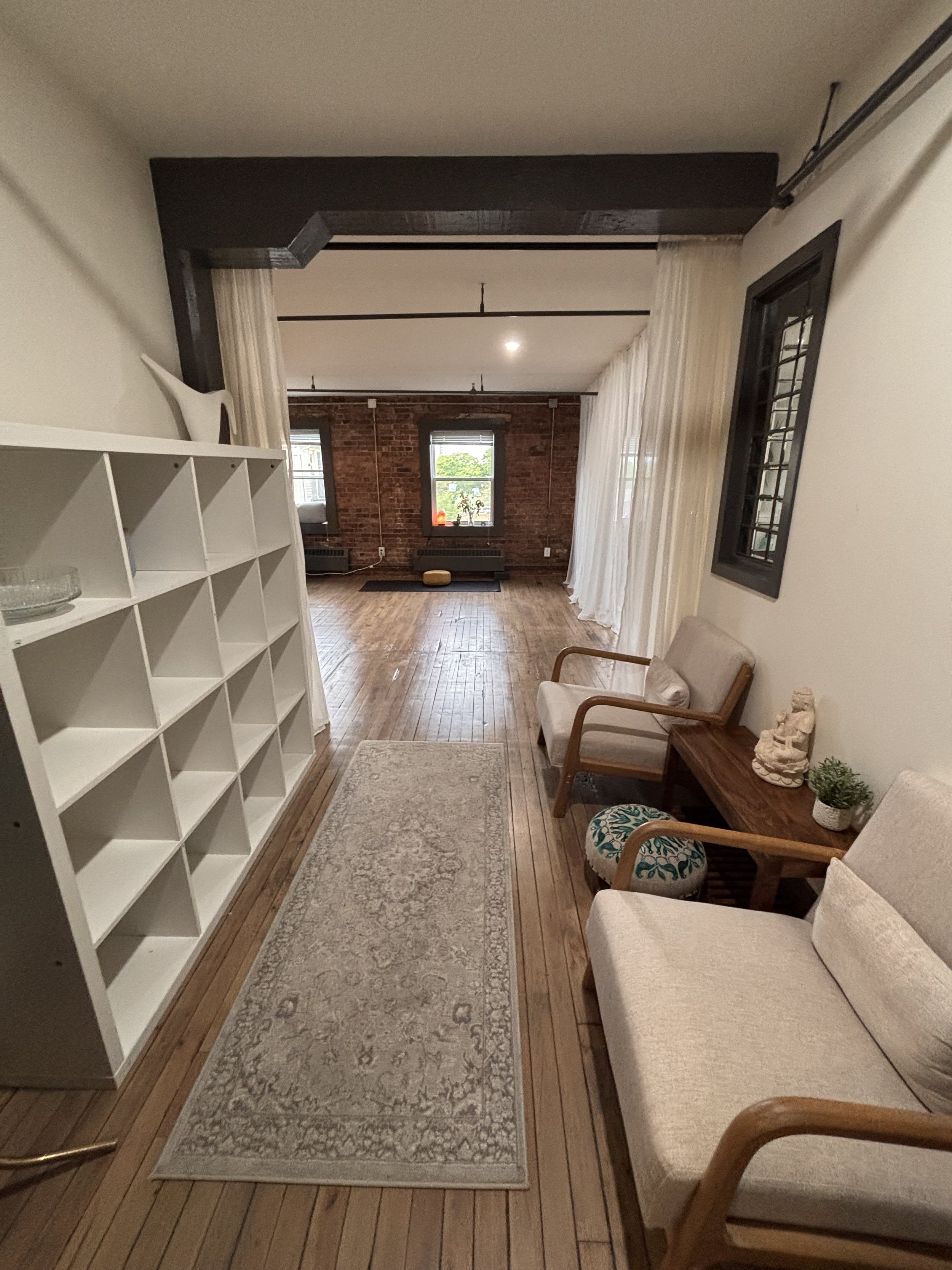 Living room with hardwood floors, a rug, white bookcase, armchairs, large window with curtains, brick wall, and decorative plants.