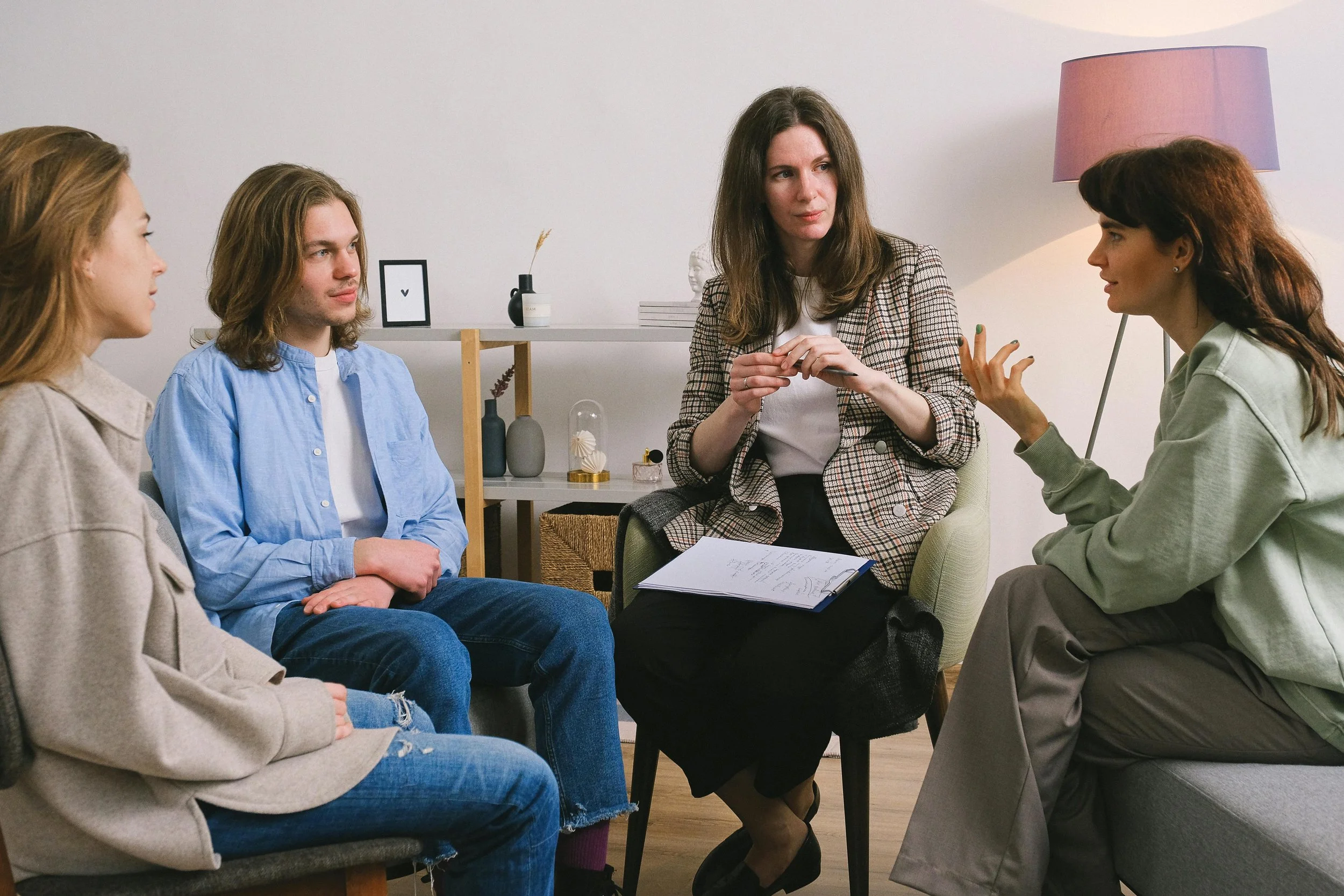 A woman with long brown hair wearing a plaid blazer is sitting and talking to four other women in a casual meeting room. The women are seated in a semicircle and appear to be engaged in a serious discussion. The background includes decorative vases, a small framed picture, and a purple lampshade.
