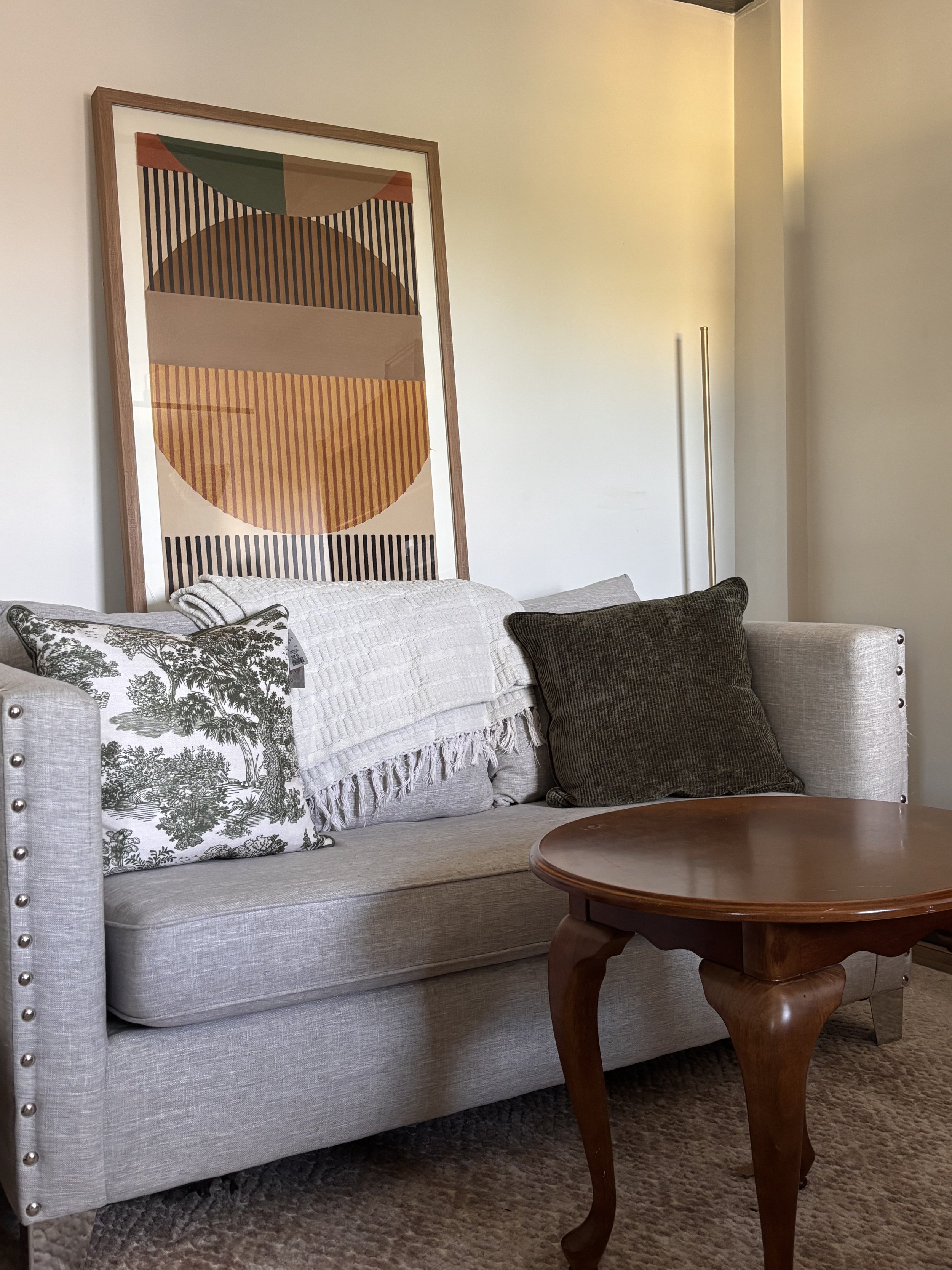 Living room corner with light-colored sofa decorated with patterned and solid pillows, a wooden coffee table, a large abstract wall art piece with geometric shapes and warm colors, and a textured area rug.