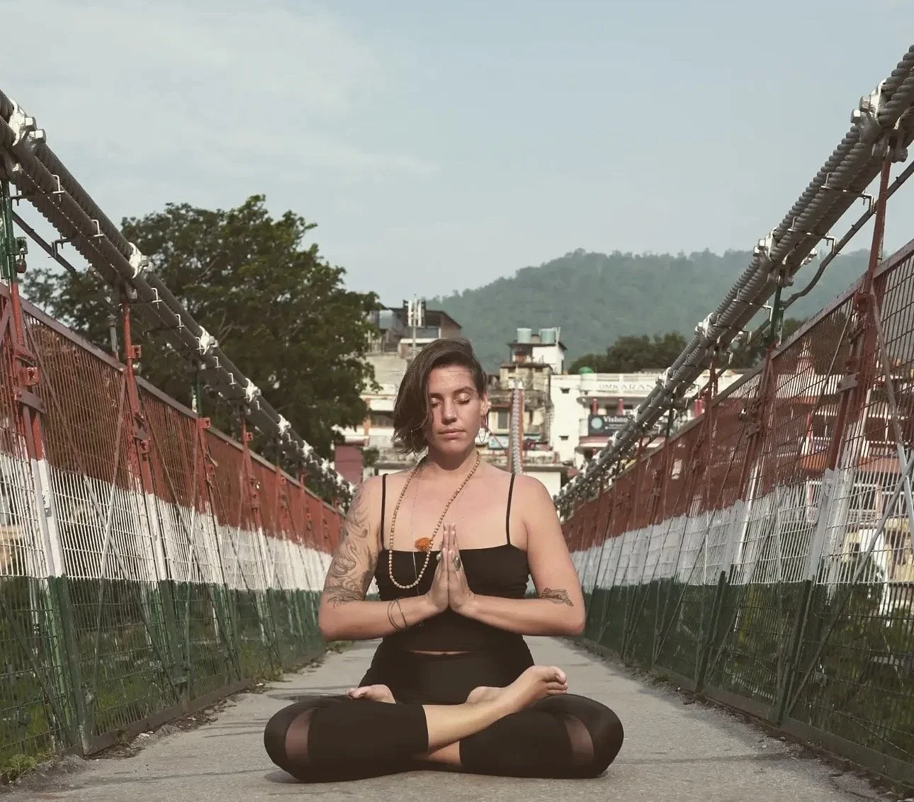 A woman practicing yoga and meditation on a bridge painted with the Indian flag colors. She is sitting in a cross-legged pose with hands in a prayer position, eyes closed, wearing a black outfit with tattoos on her arms, and jewelry around her neck. Hills and buildings are visible in the background.