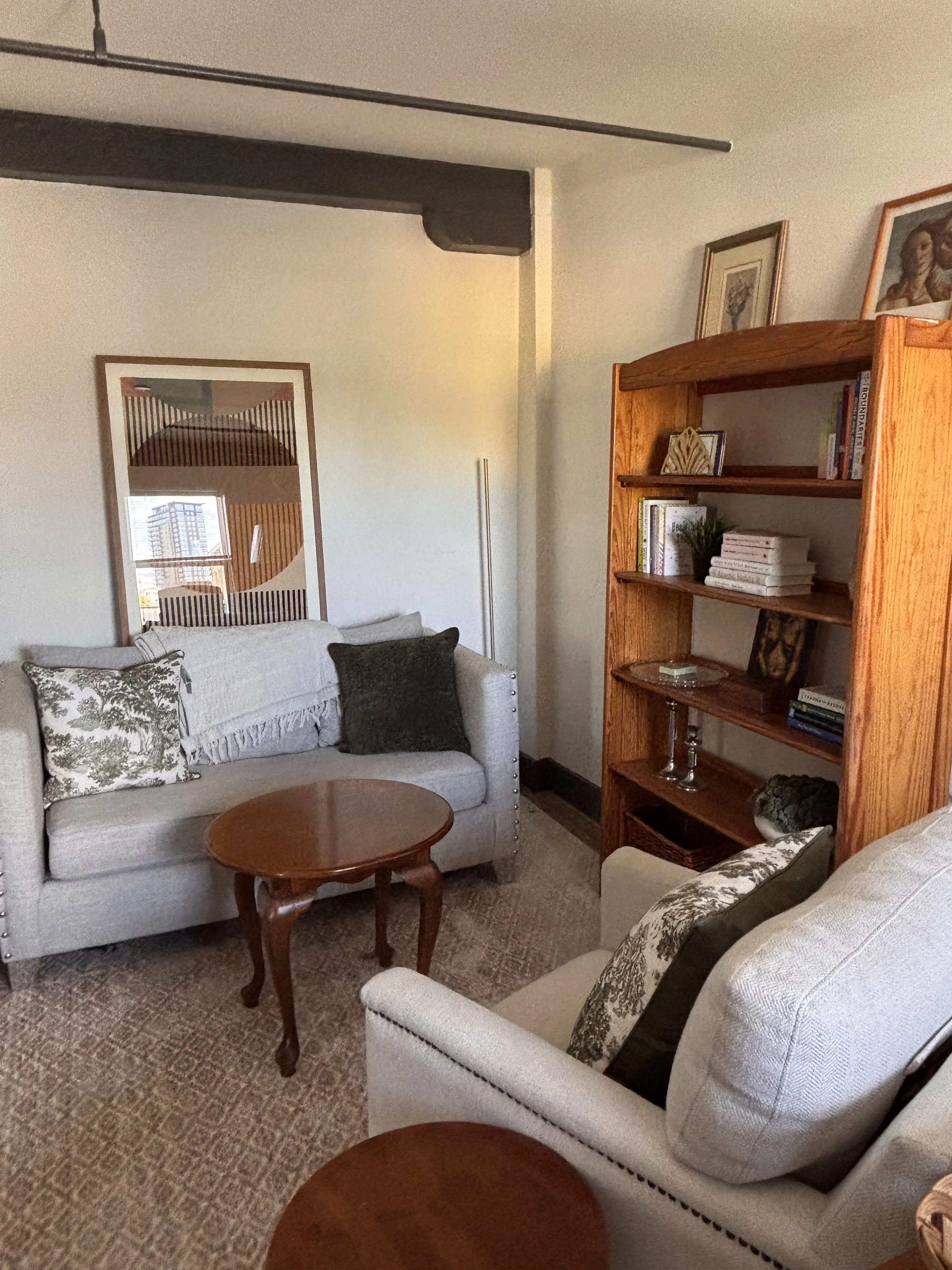 A cozy living room with two white upholstered sofas, patterned throw pillows, a wooden coffee table, a wooden bookshelf with books and decorative items, framed artwork on the walls, and a large window with a city view.