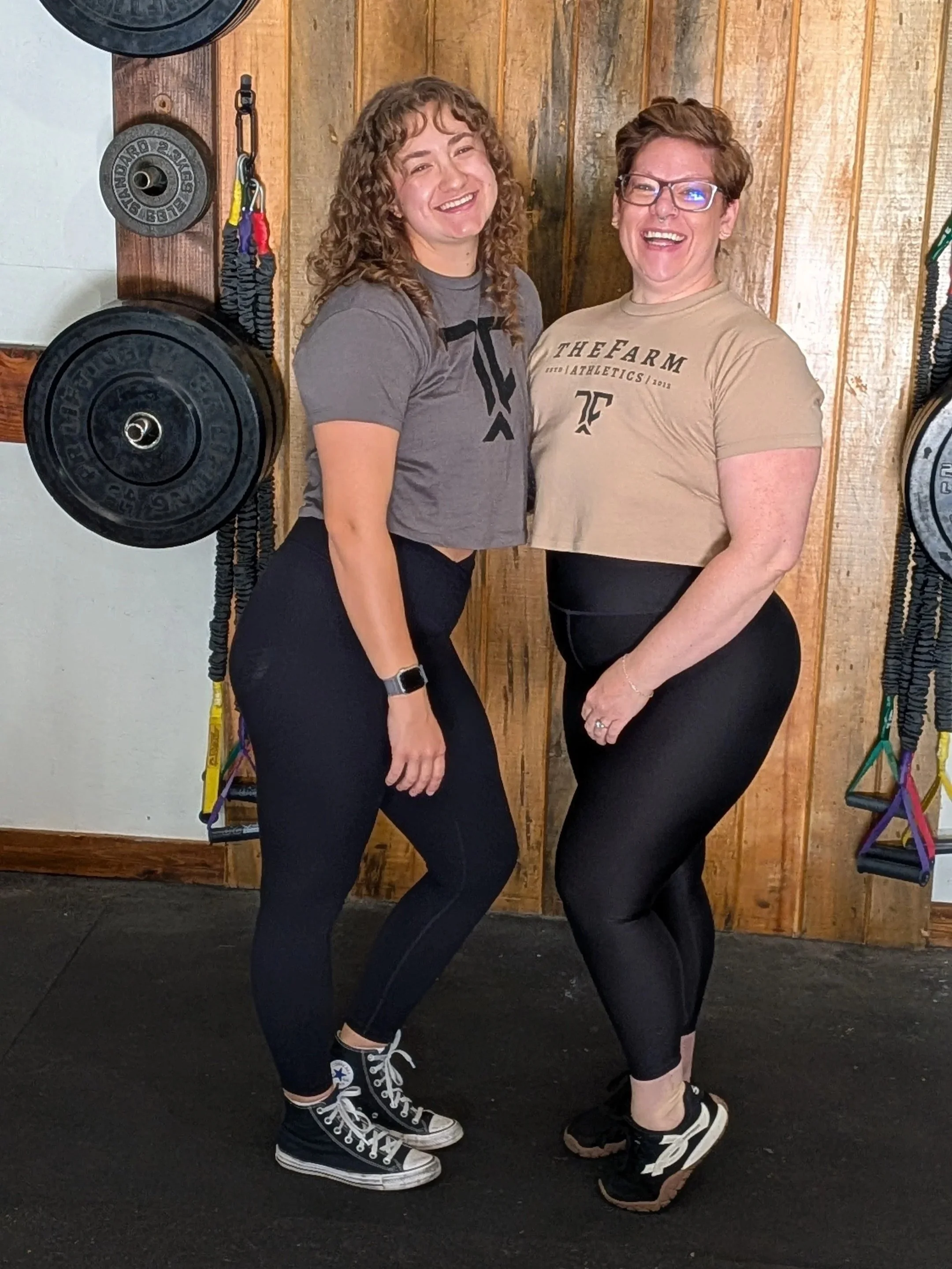 Two women smiling and posing together in a gym with wooden walls and fitness equipment, including weight plates and resistance bands, behind them.