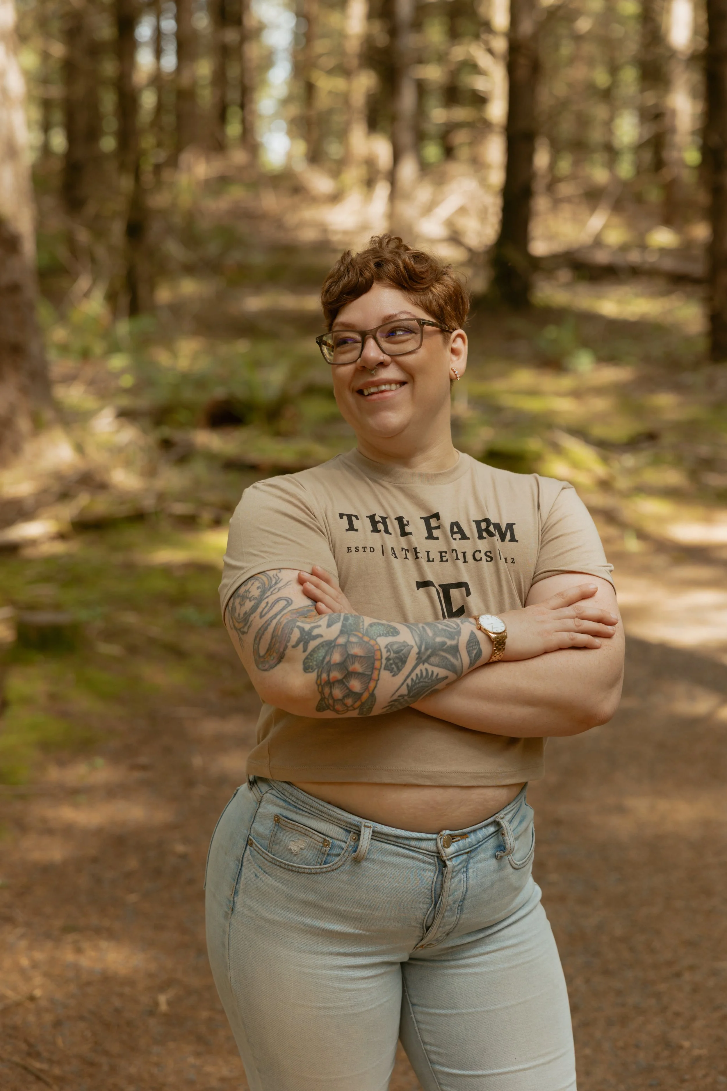 A woman with short curly hair and glasses is standing in a forest, smiling and crossing her arms. She has tattoos on her left arm and is wearing a tan T-shirt with