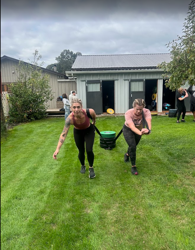 Two women are doing tug-of-war with a black strap attached to a green sled on a grassy backyard. In the background, there are other people near a small shed and garden.