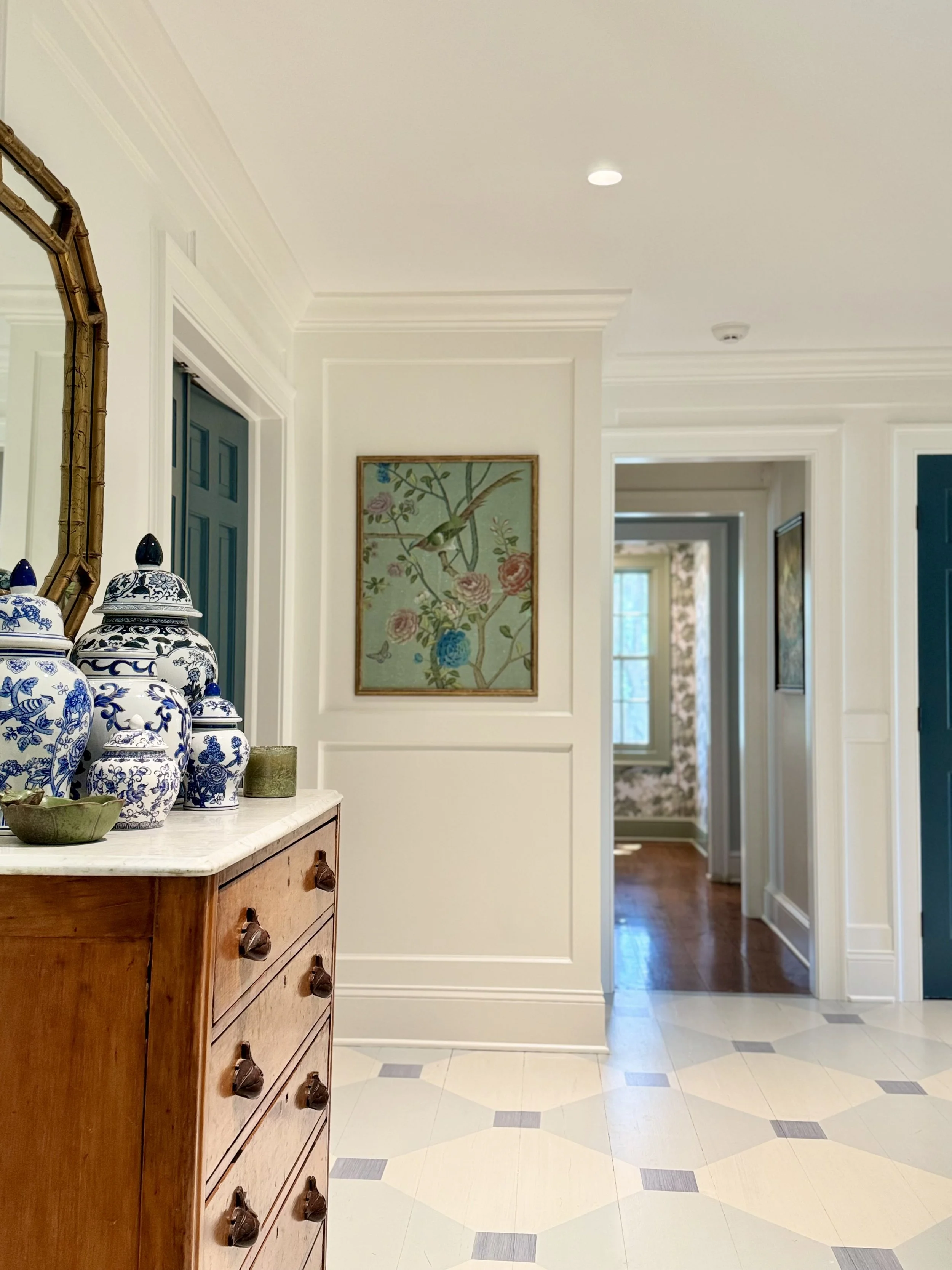 Entryway with a wooden dresser topped with blue and white ceramic jars, a mirror on the wall, and a hallway leading to rooms with floral wallpaper and hardwood floors