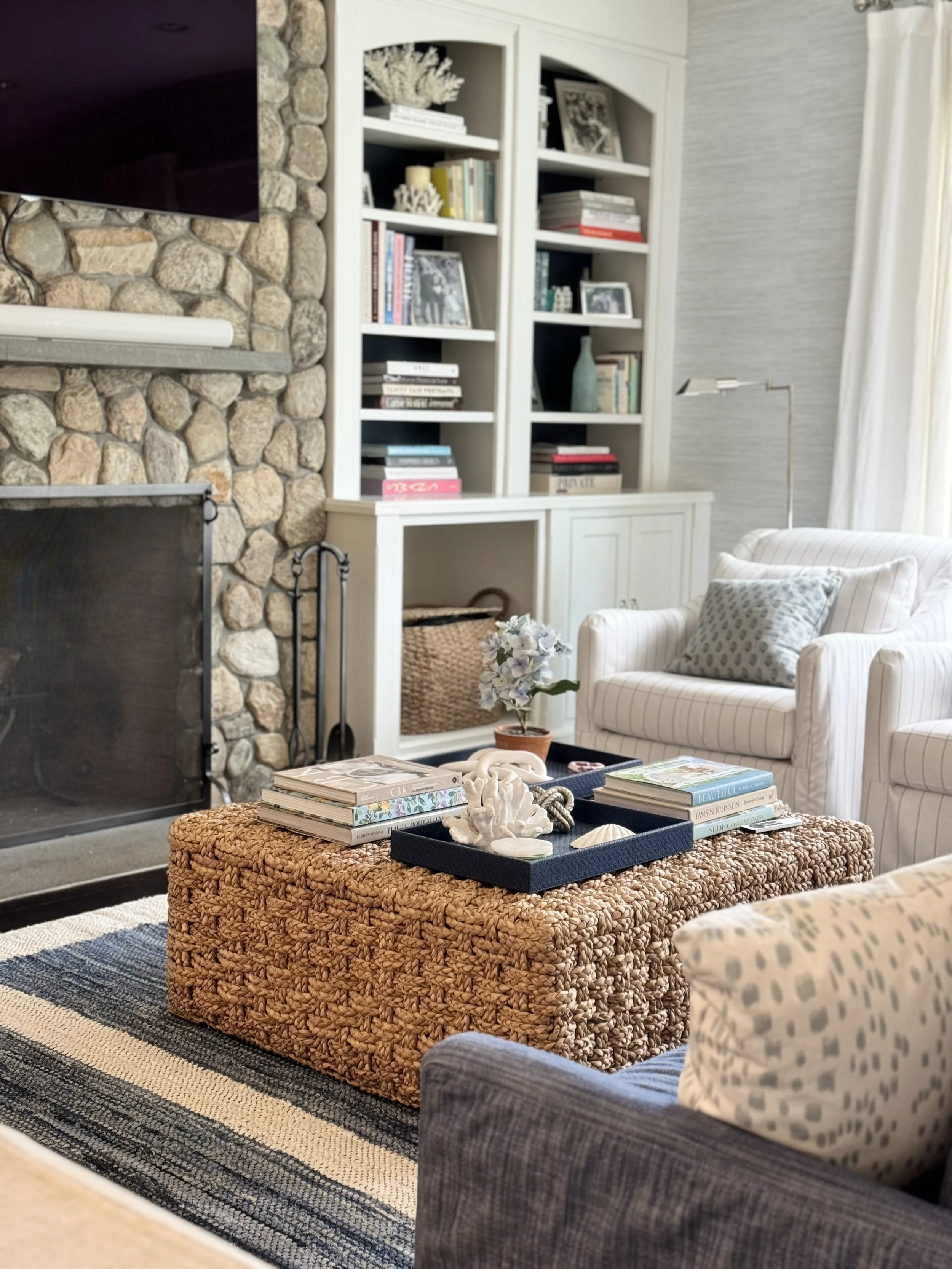 Cozy living room with stone fireplace, white built-in shelves filled with books and decor, a woven ottoman with seashells and books on top, a small potted plant, and white upholstered chairs with patterned pillows.