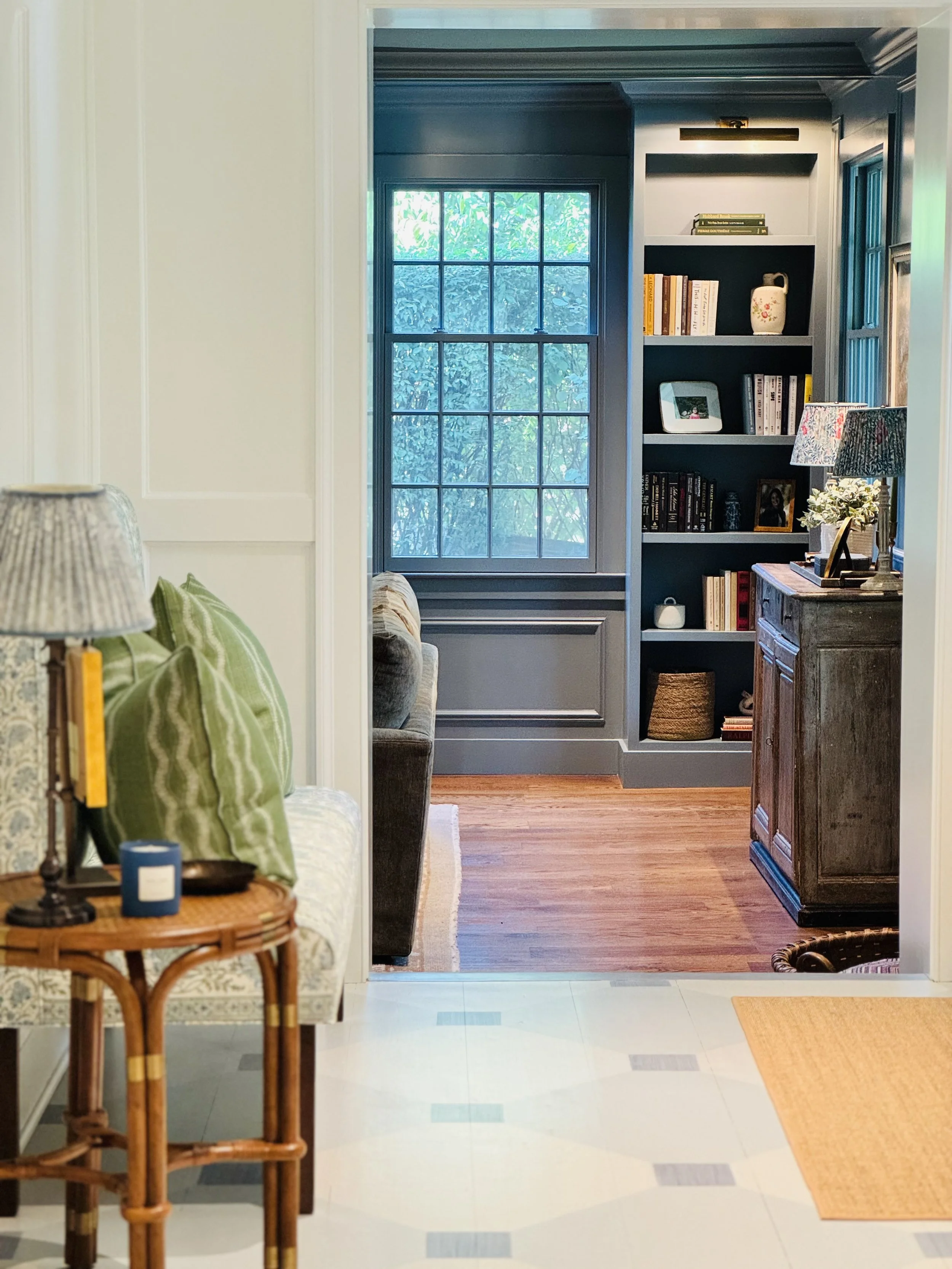 Interior view of a home with a doorway leading to a room with hardwood floors, a large window, and built-in bookshelves filled with books, decorative items, and framed photos, with a sideboard and lamps in the foreground.