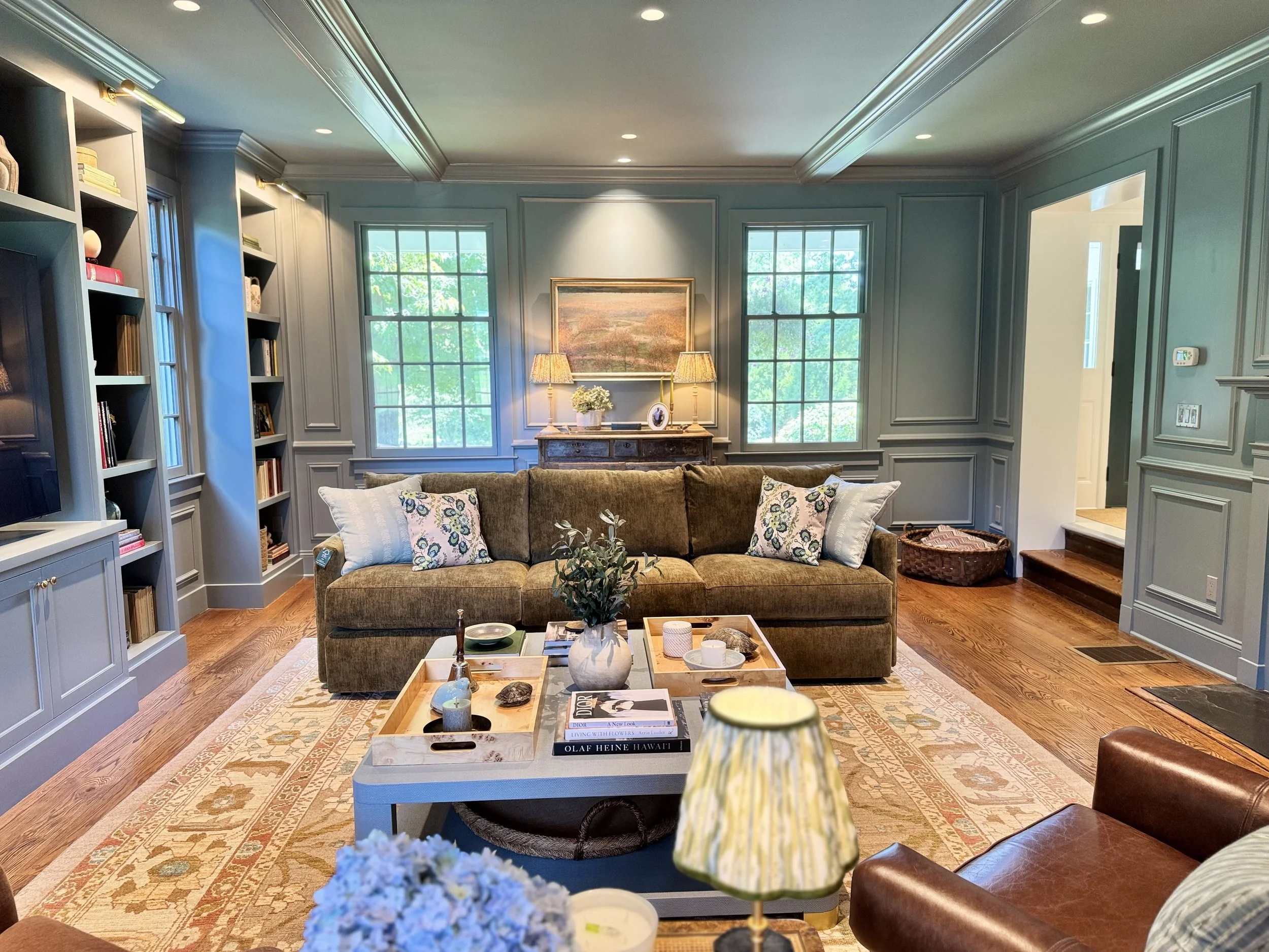 A cozy living room with a green sofa, decorative pillows, a wooden coffee table with trays and books, built-in bookshelves, three windows with greenery outside, a side cabinet with lamps and a painting, a patterned rug, a leather armchair, and warm lighting.