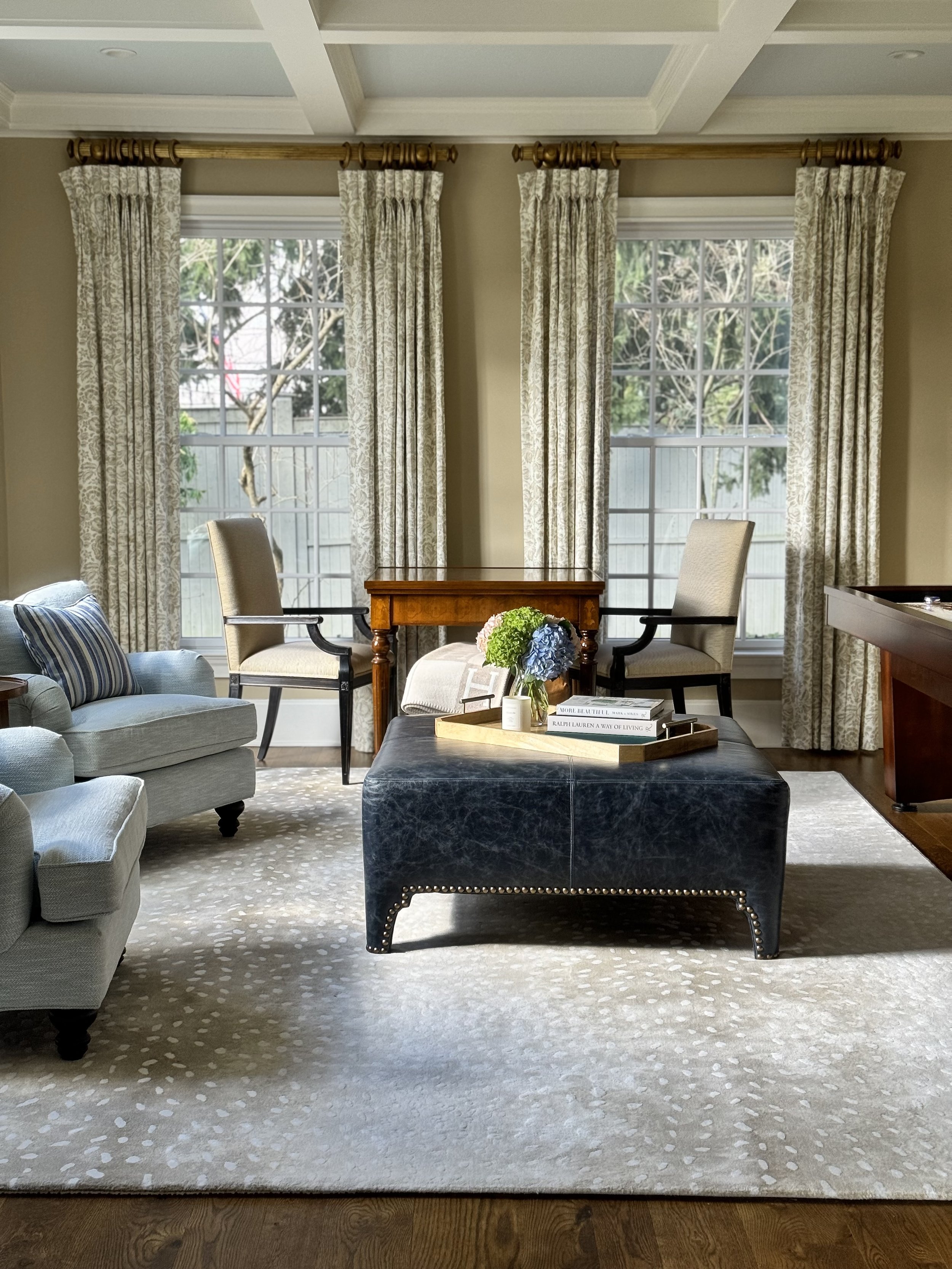 Living room featuring large windows with patterned curtains, beige walls, a black leather ottoman with studded details, a tray with flowers and books, an upholstered armchair, a loveseat with striped pillows, and a wooden table.