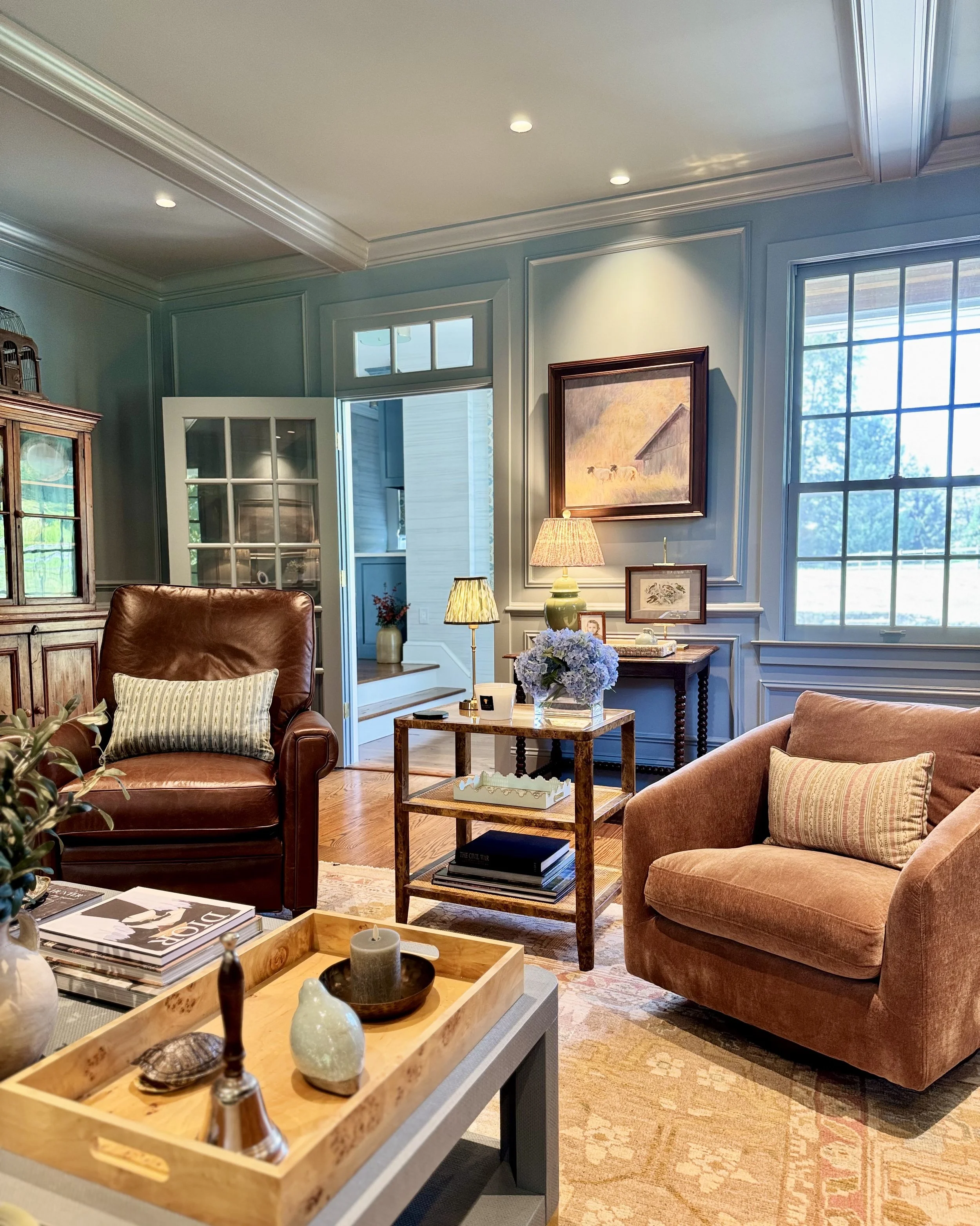 Living room with blue walls, large window, brown leather armchair, pink velvet armchair, wooden side table with flowers and lamps, framed pictures on wall, decorative tray with candles on coffee table, hardwood floor, rug.
