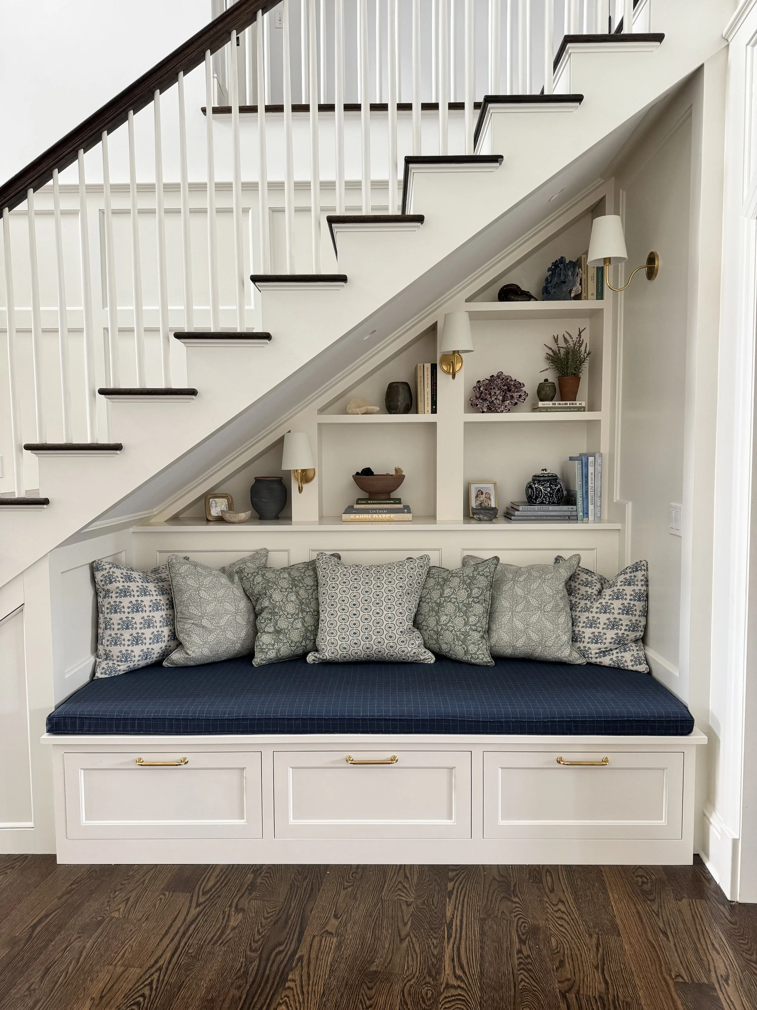 Built-in corner seat beneath a staircase with white cabinetry, a cushioned navy blue seat, and a row of decorative pillows. Above the seat, built-in shelves hold books, vases, rocks, and decorative objects, with three small wall-mounted lamps providing lighting.