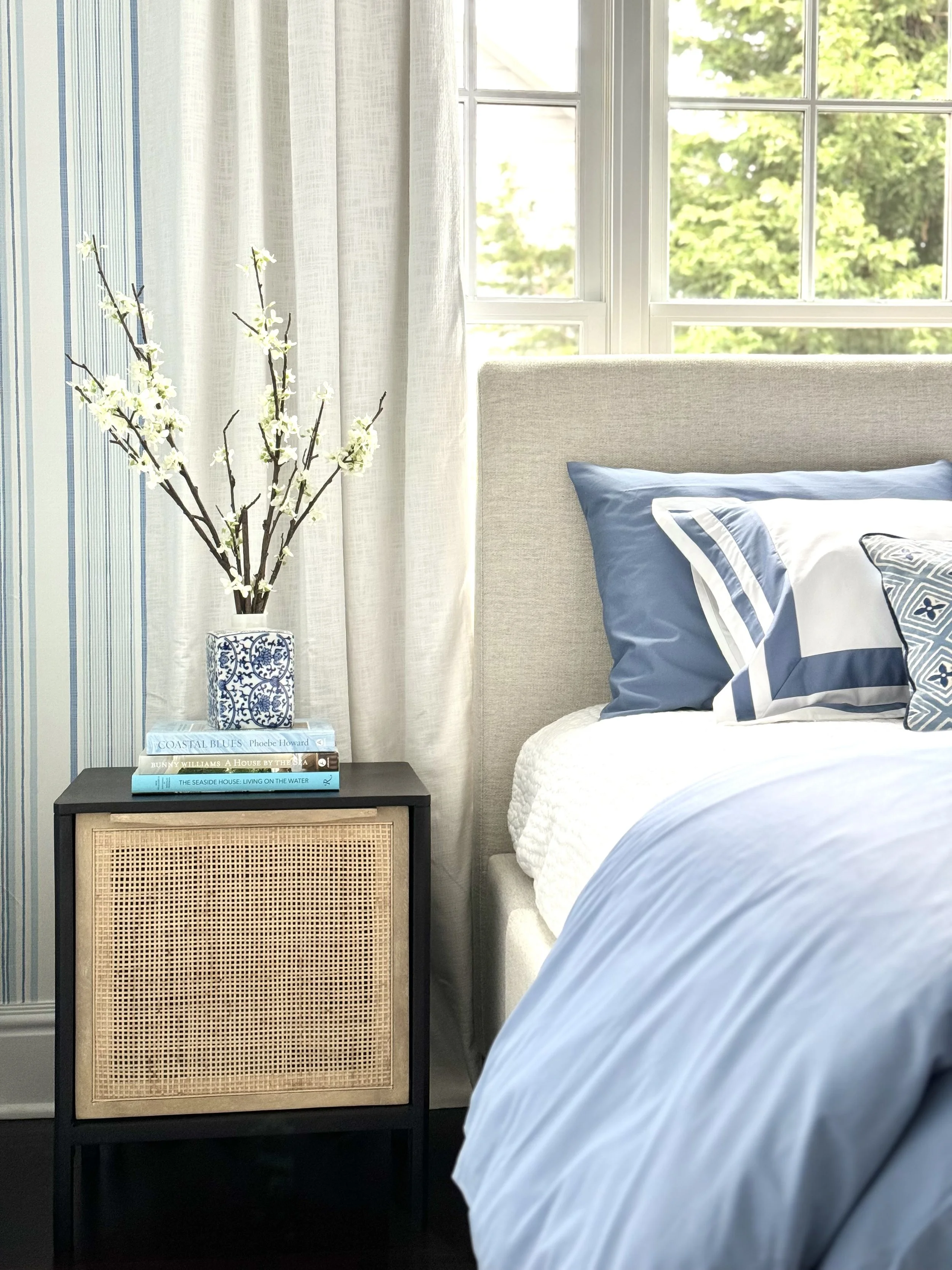 Nightstand with a vase of white flowering branches, a stack of books, next to a bed with blue and white bedding in a bright room with large windows and white curtains.
