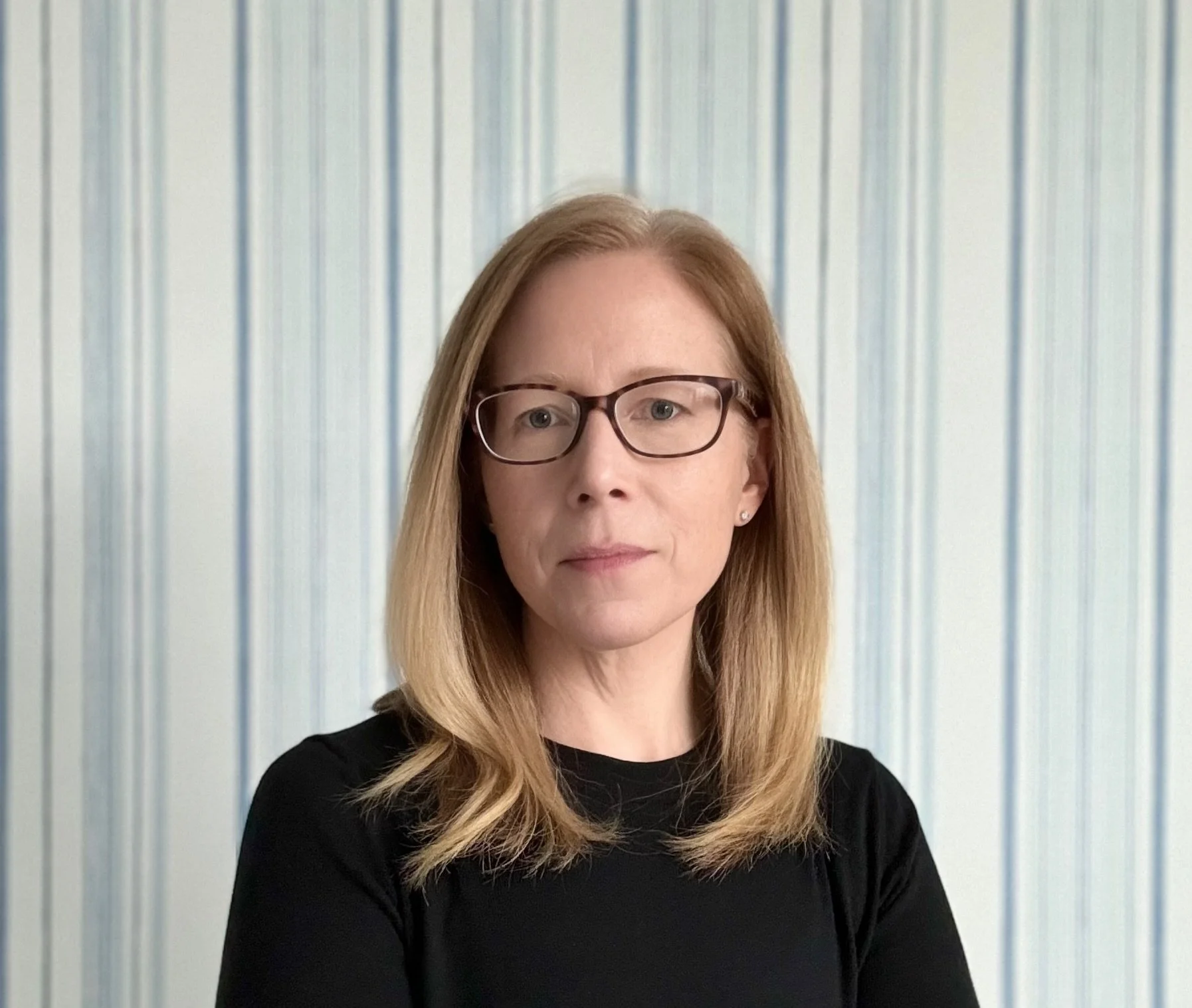 Woman with shoulder-length red hair and glasses wearing a black top posing in front of a blue and white striped background.