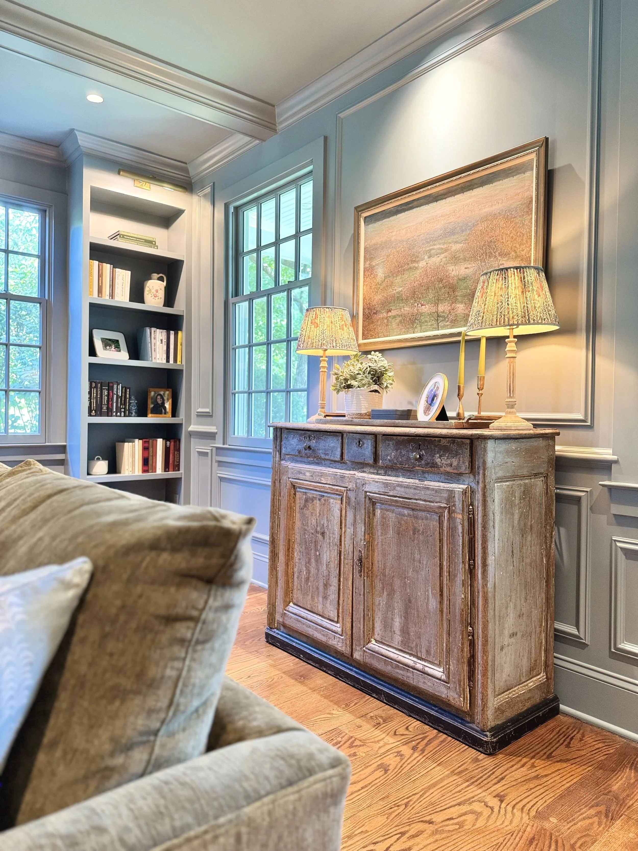 A cozy living room corner with a beige armchair, a wooden sideboard, two table lamps, a framed painting, a potted plant, and a bookshelf in the background near windows.