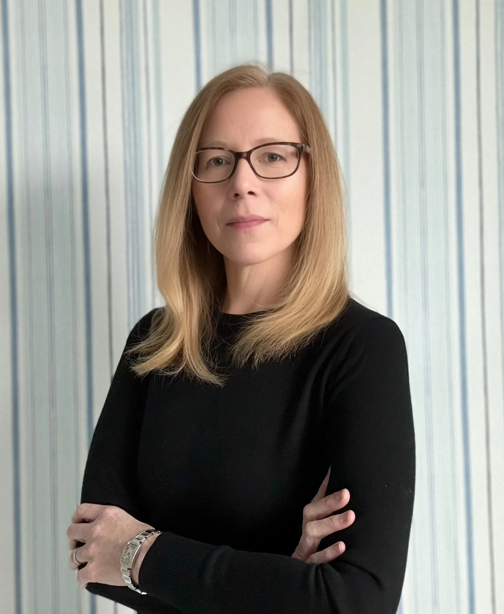 A woman with blonde hair, glasses, and a black long-sleeve top standing with arms crossed in front of a striped blue and white background.