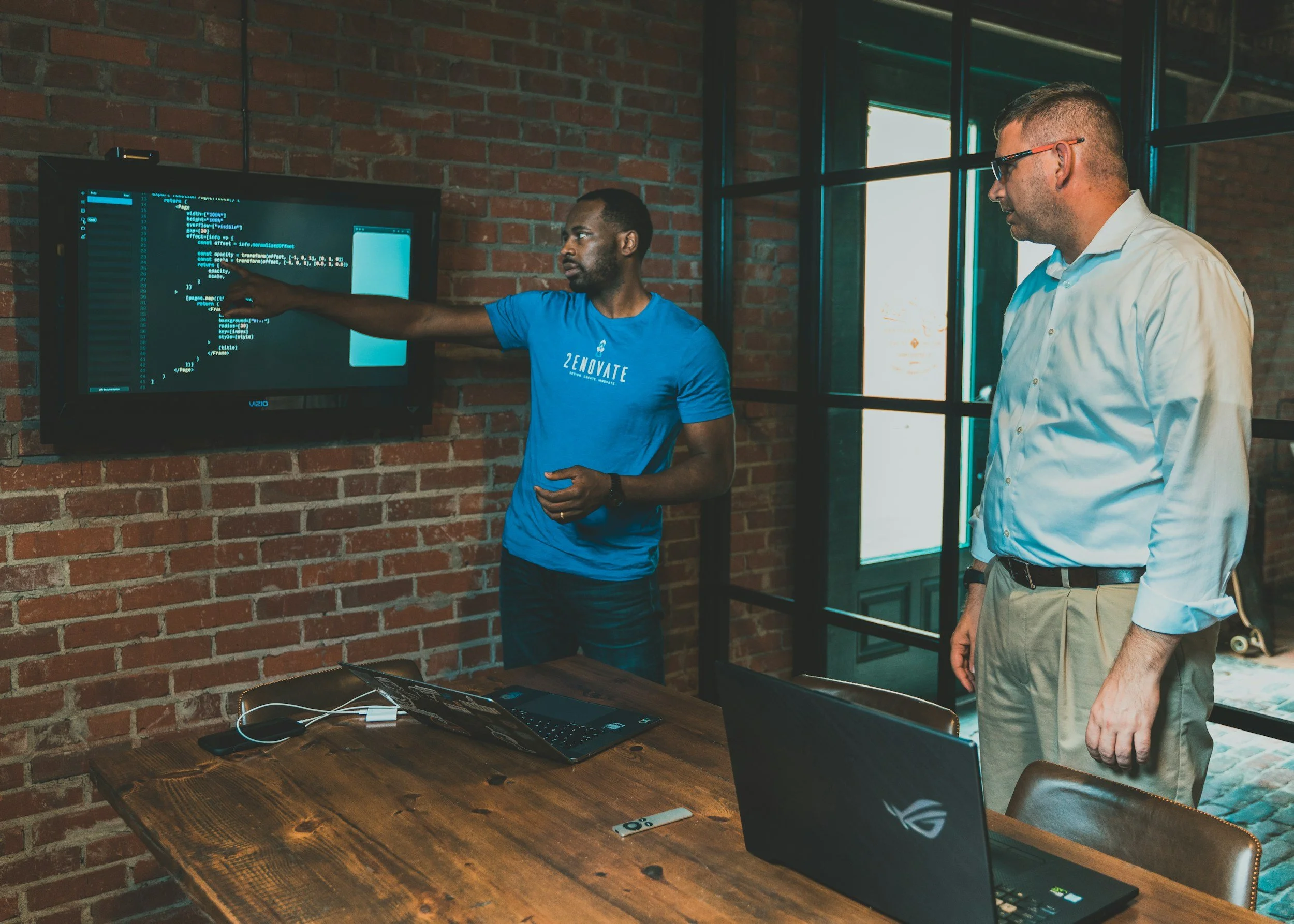 Two men in a modern office with exposed brick walls; one is presenting code on a large screen, the other is listening. A laptop and a remote are on the wooden table.