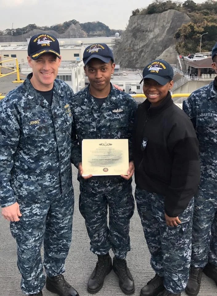 Three sailors in navy camouflage uniforms and hats, standing outdoors on a dock, some holding an award certificate, with a rocky hillside and water in the background.