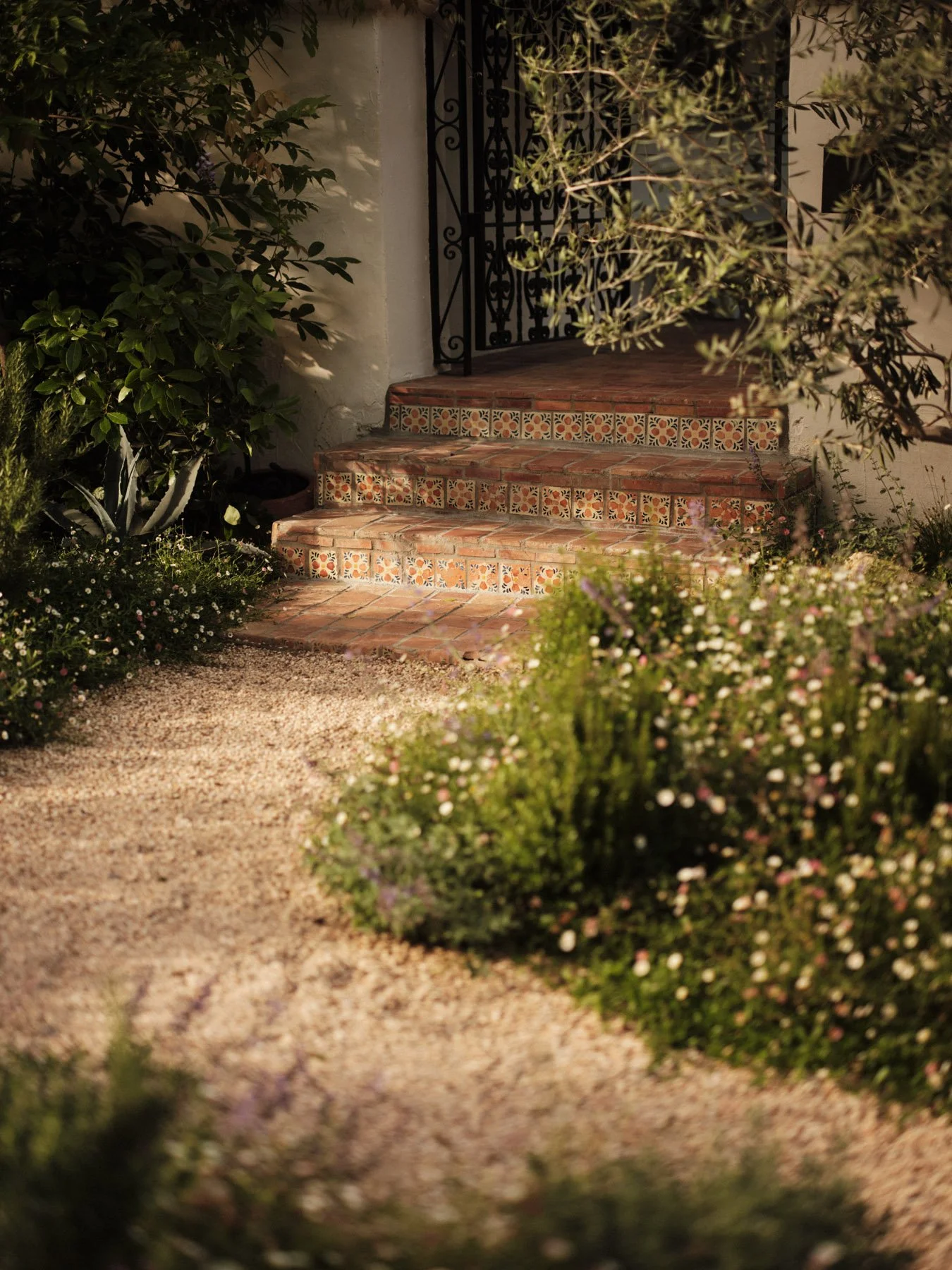 A set of brick and tile steps leading up to a house entrance, surrounded by greenery and small flowering plants, with an iron gate at the top of the steps.