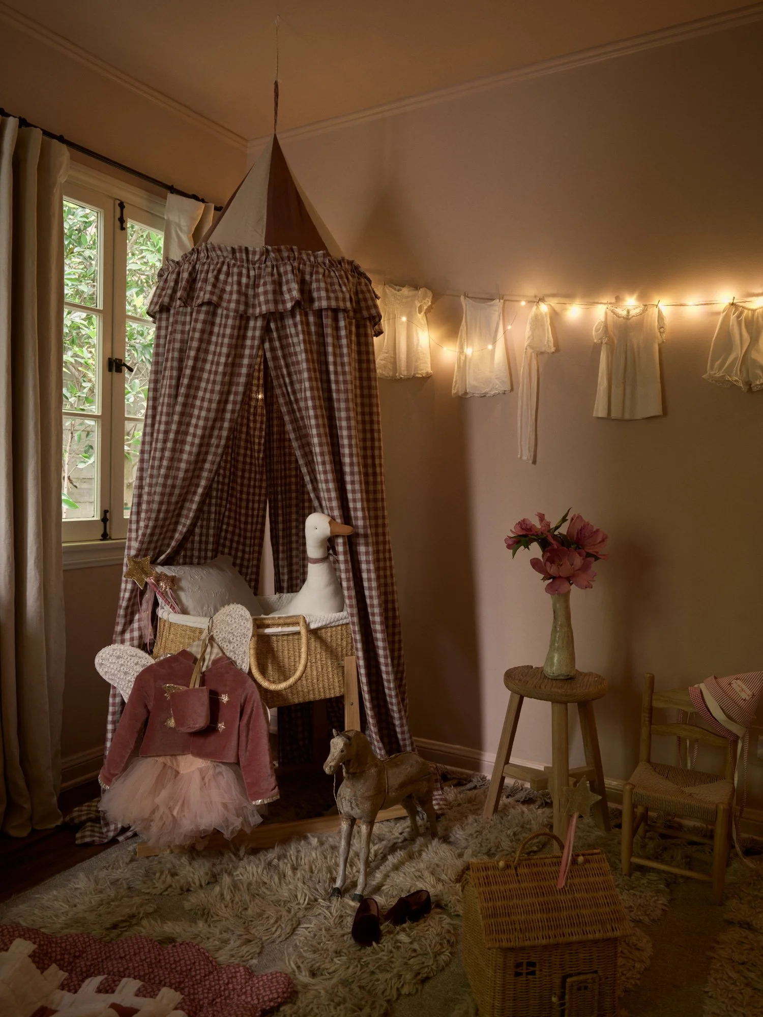 A cozy children's room with a pink and white checked canopy tent, fairy string lights, and a stuffed duck in the basket. Decorative clothing items hang on a string across the wall, and there are toys including a wooden horse, shoes, and a small chair