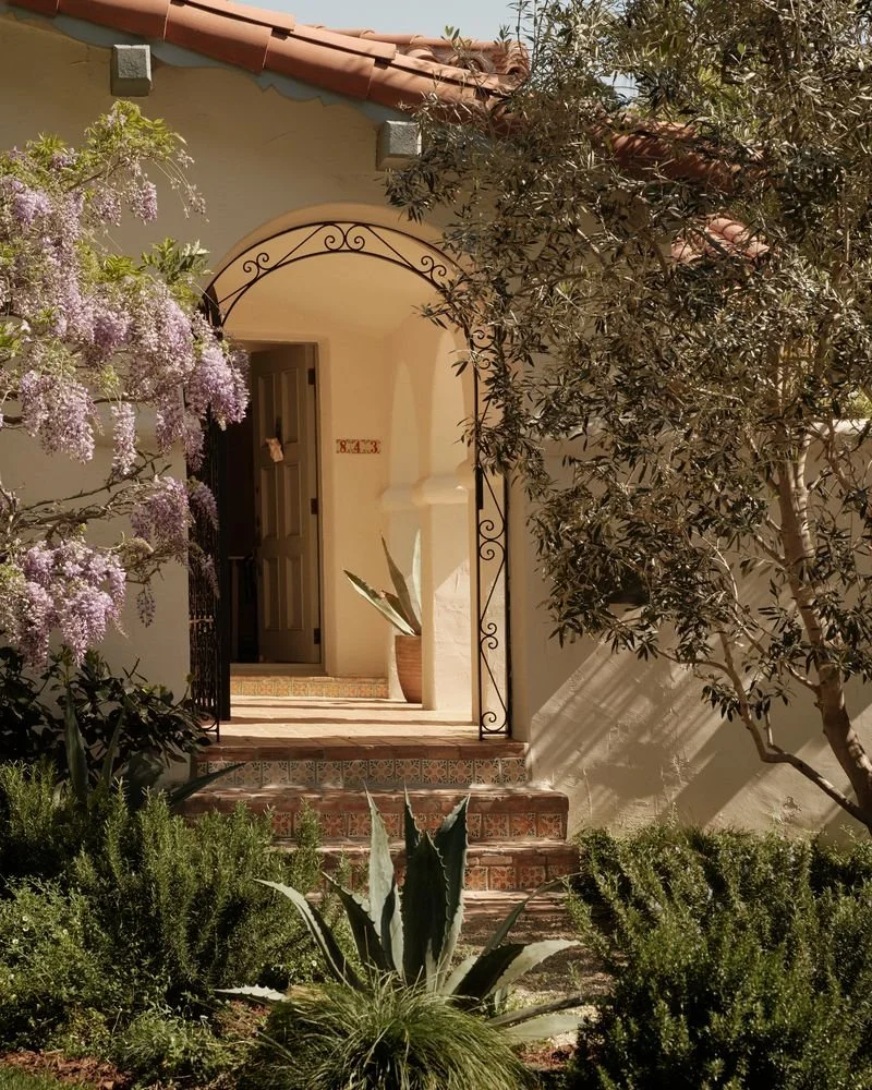 Front entrance of a house with a tiled staircase, potted plants, flowering purple and green bushes, and trees, under a tiled roof with a decorative iron archway.