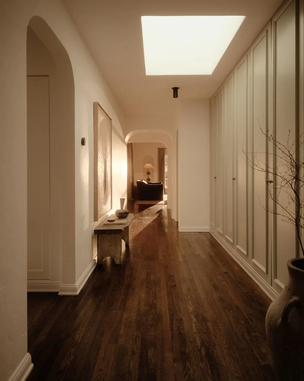 A hallway with wooden flooring, white walls, and built-in white cabinets on the right. There is a small stone table with bowls on it and artwork on the wall. The hallway leads to a seating area with a lamp and armchair in the background, illuminated 