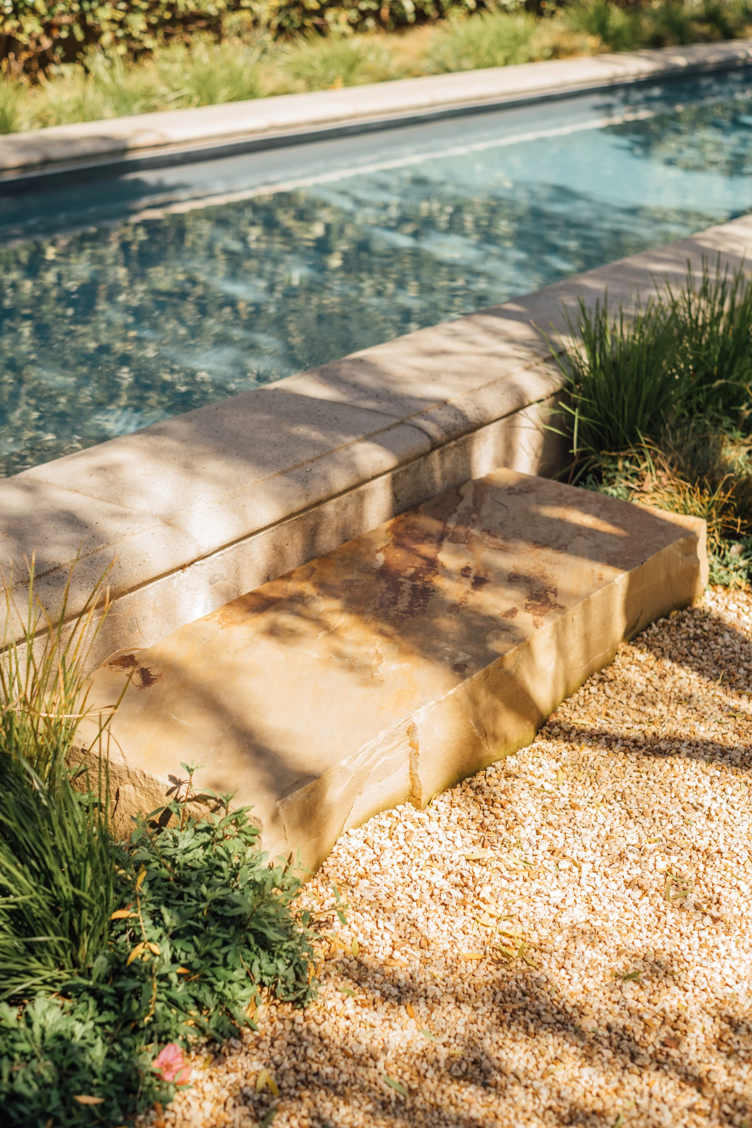 Close-up of a crystal-clear swimming pool with a stone edge, surrounded by gravel and plants, in warm sunlight.