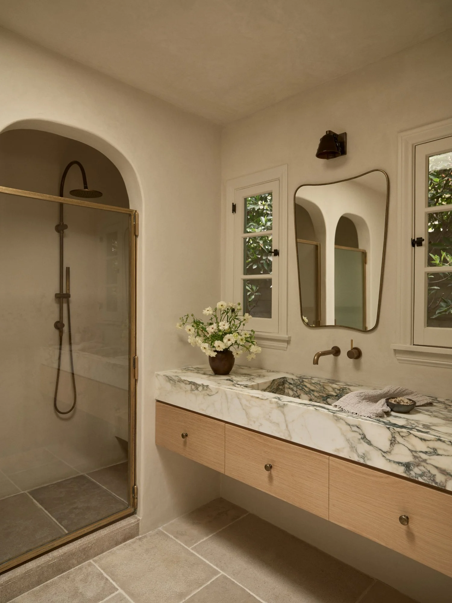 Modern bathroom with a marble countertop, oval mirror, wooden vanity, flower vase, and walk-in shower with glass door and brass hardware.