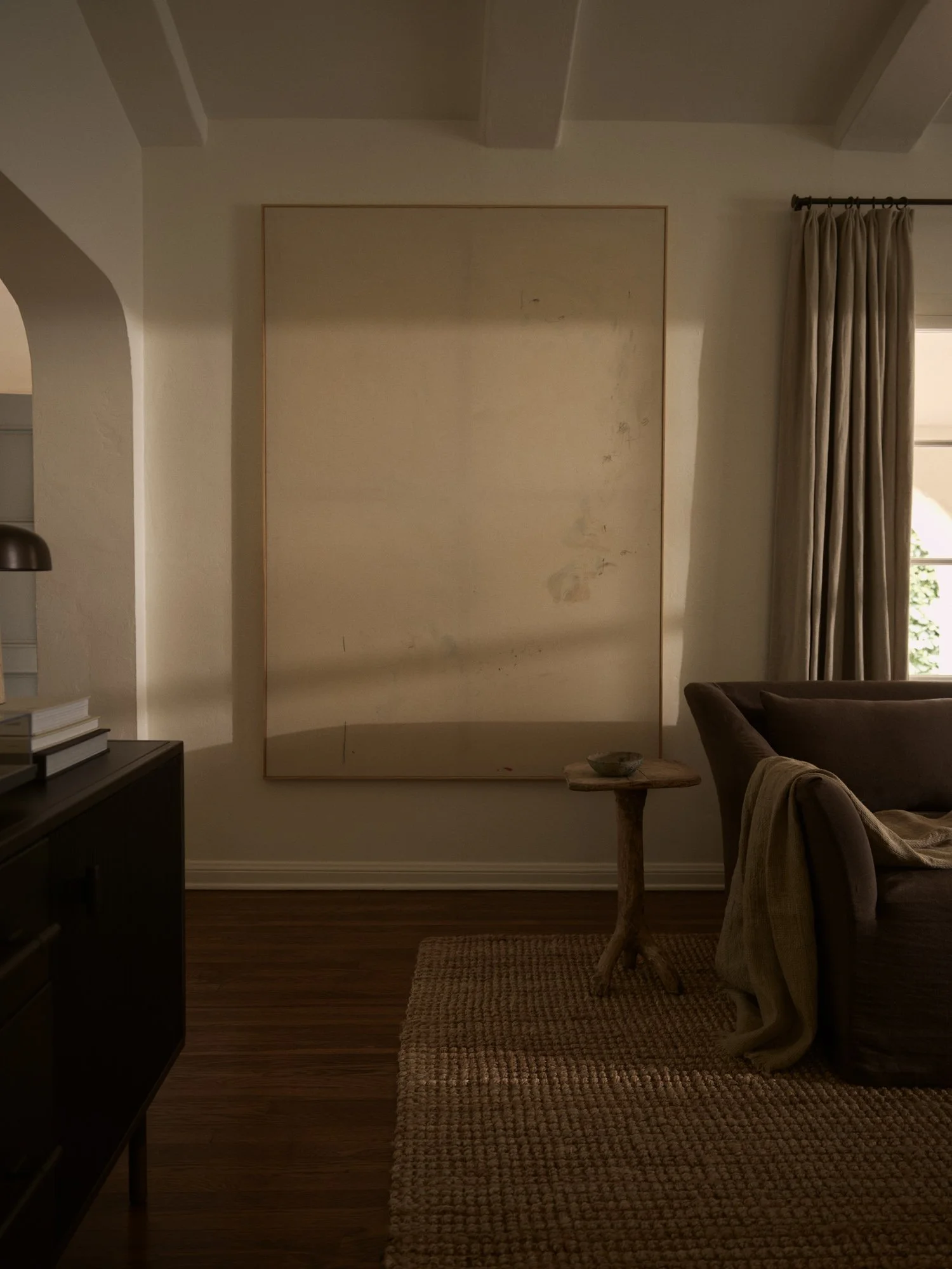 Living room with a large mirror on the wall, beige curtains, a brown sofa with a throw blanket, a small rustic side table, wooden flooring, and a woven rug.