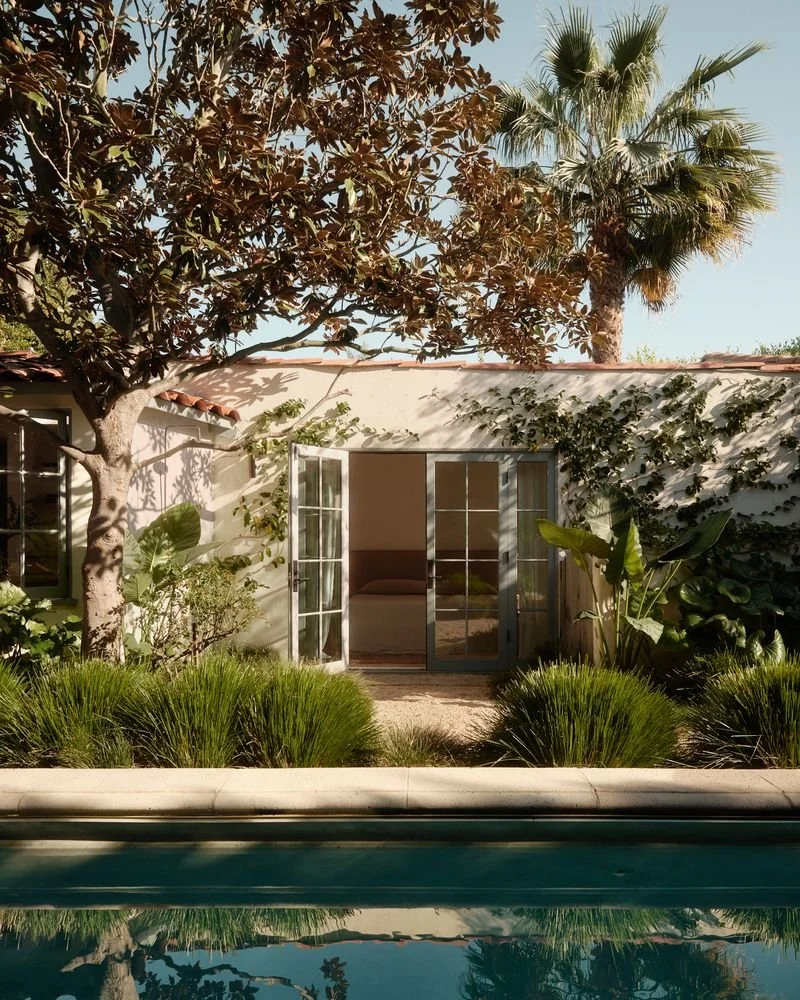 A backyard scene with a swimming pool in the foreground, lush green bushes, a large tree, and a tropical palm tree. In the background, there is a sliding glass door leading into a house with a small patio area.