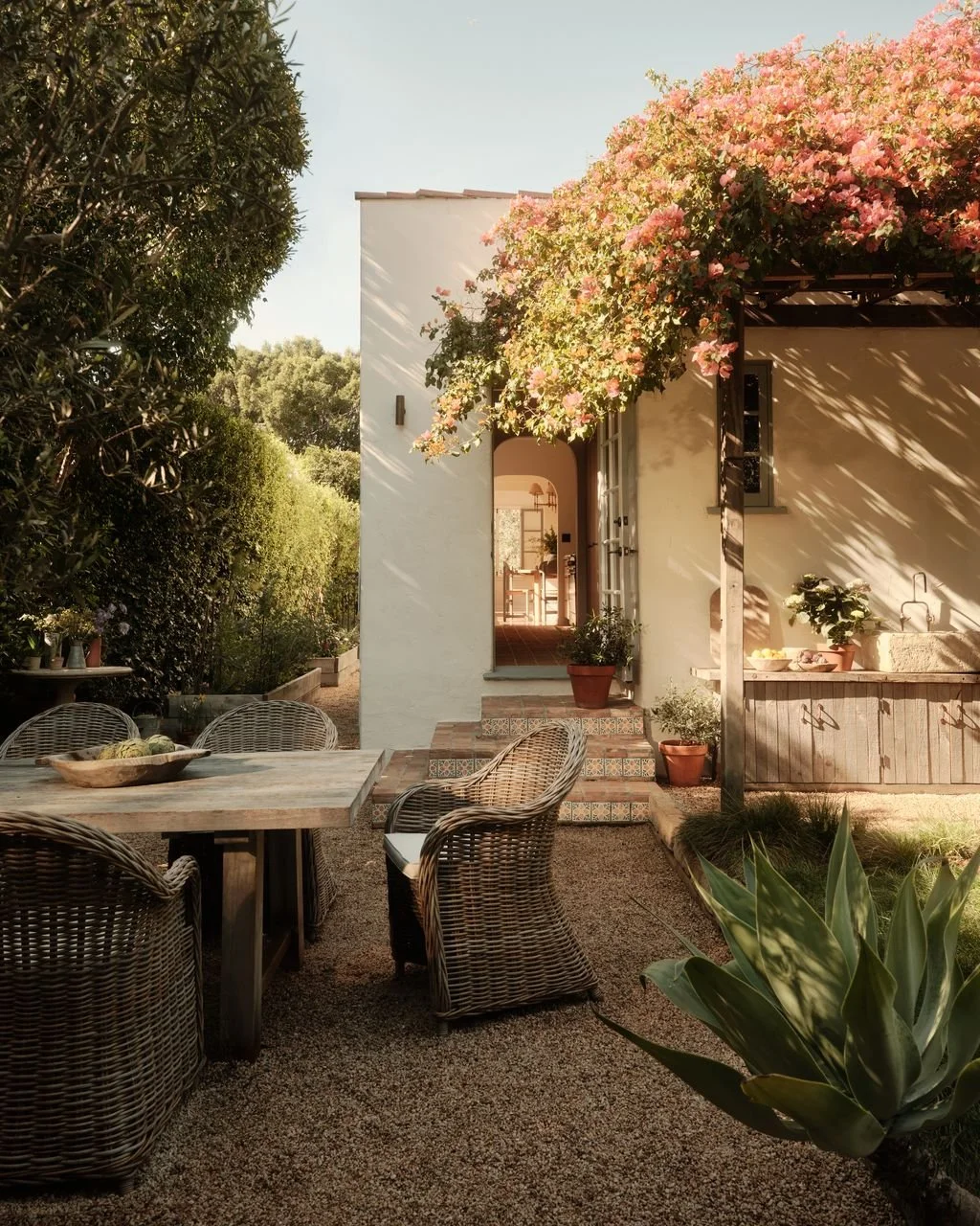 A cozy outdoor patio with a wooden table and wicker chairs in front of a white house with stairs, potted plants, and a flowering pink bougainvillea tree, surrounded by lush green plants and trees.