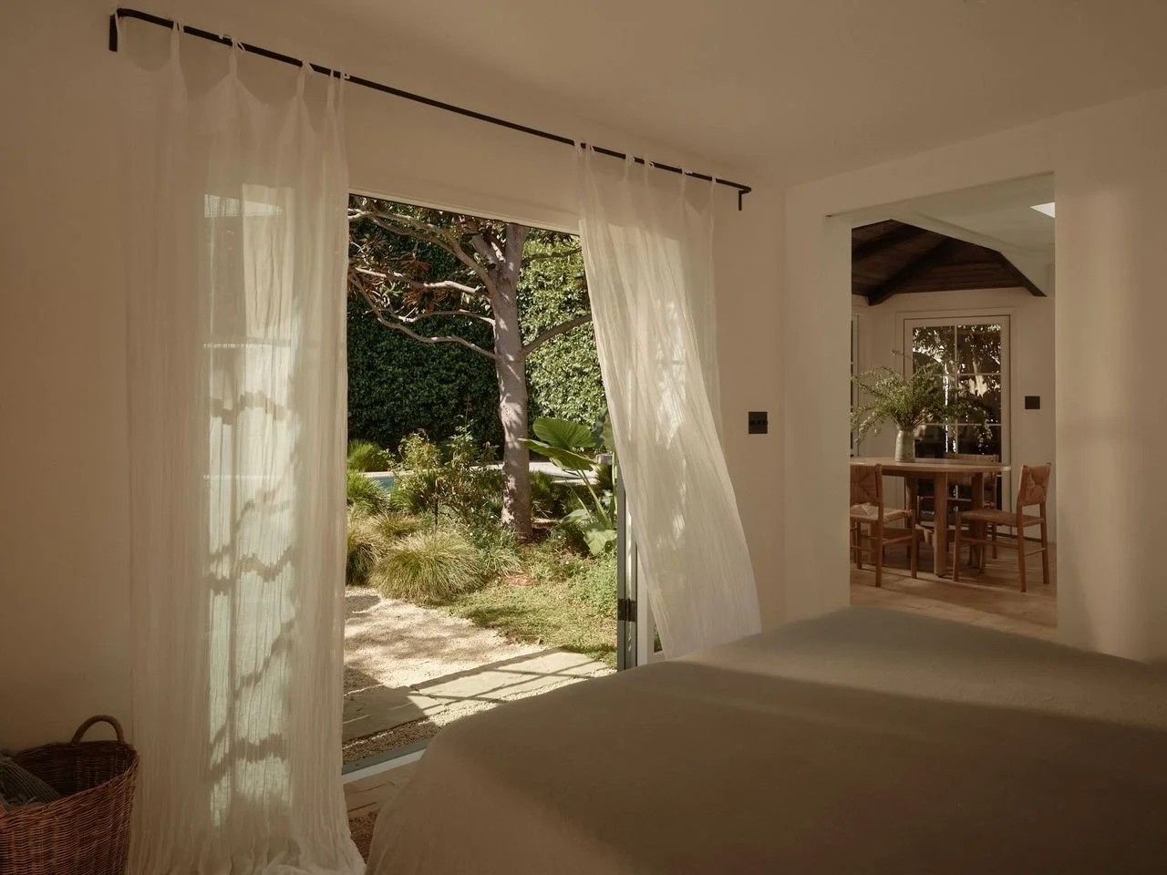 A bedroom with an open sliding door showing a garden with trees and plants outside. Light, sheer curtains hang over the door. Inside, part of a bed and a basket are visible. In the background, a dining area with a wooden table, chairs, and a vase wit
