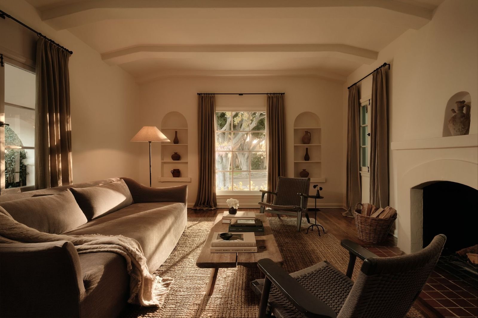 Cozy living room with beige sofa, wooden coffee table, armchair, fireplace, and large window with beige curtains, decorated with vases and a floor lamp.