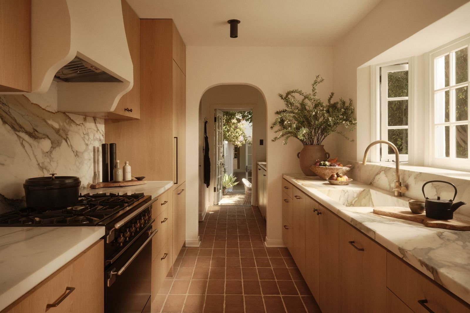 Sunlit farmhouse kitchen with wooden cabinets, a marble countertop, a black kettle on the sink, and decorative bowls and plants near the window.