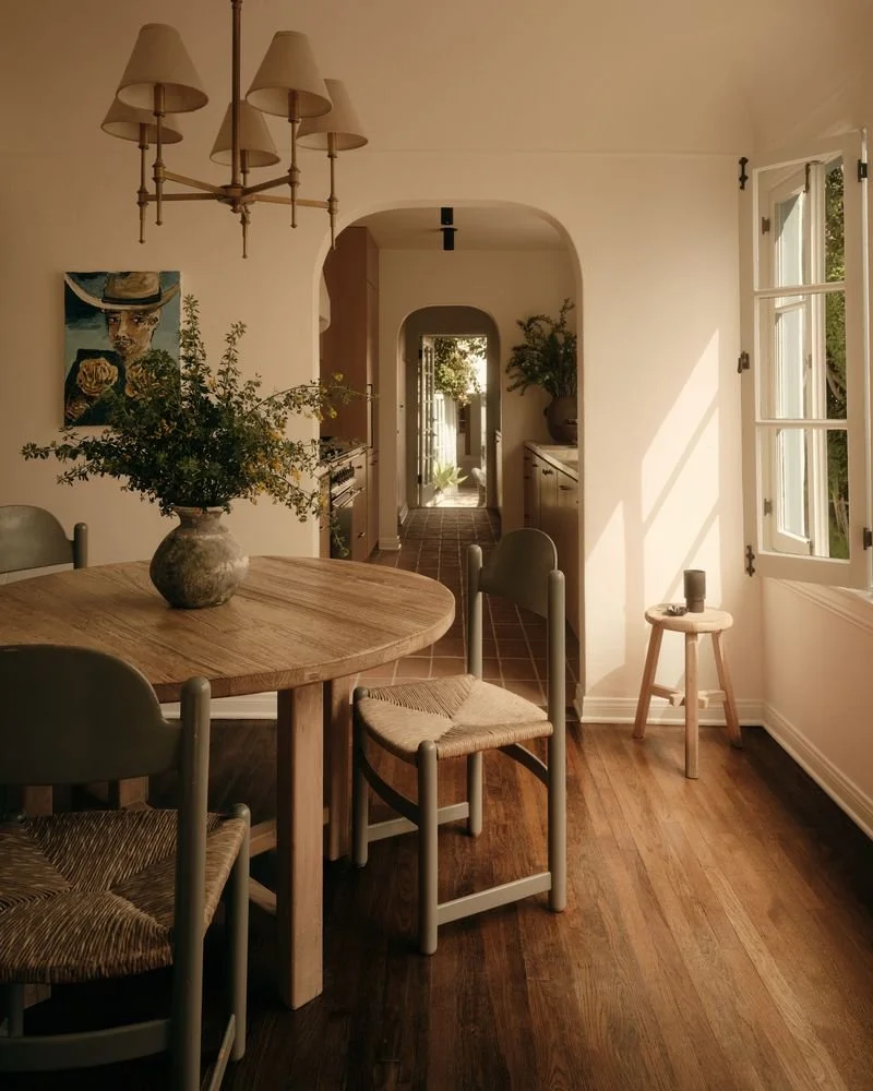 A dining area with a round wooden table, a vase of flowers, four chairs with woven seats, a doorway leading to a hallway, open window with sunlight, a small stool with a candle, and ceiling light fixtures.